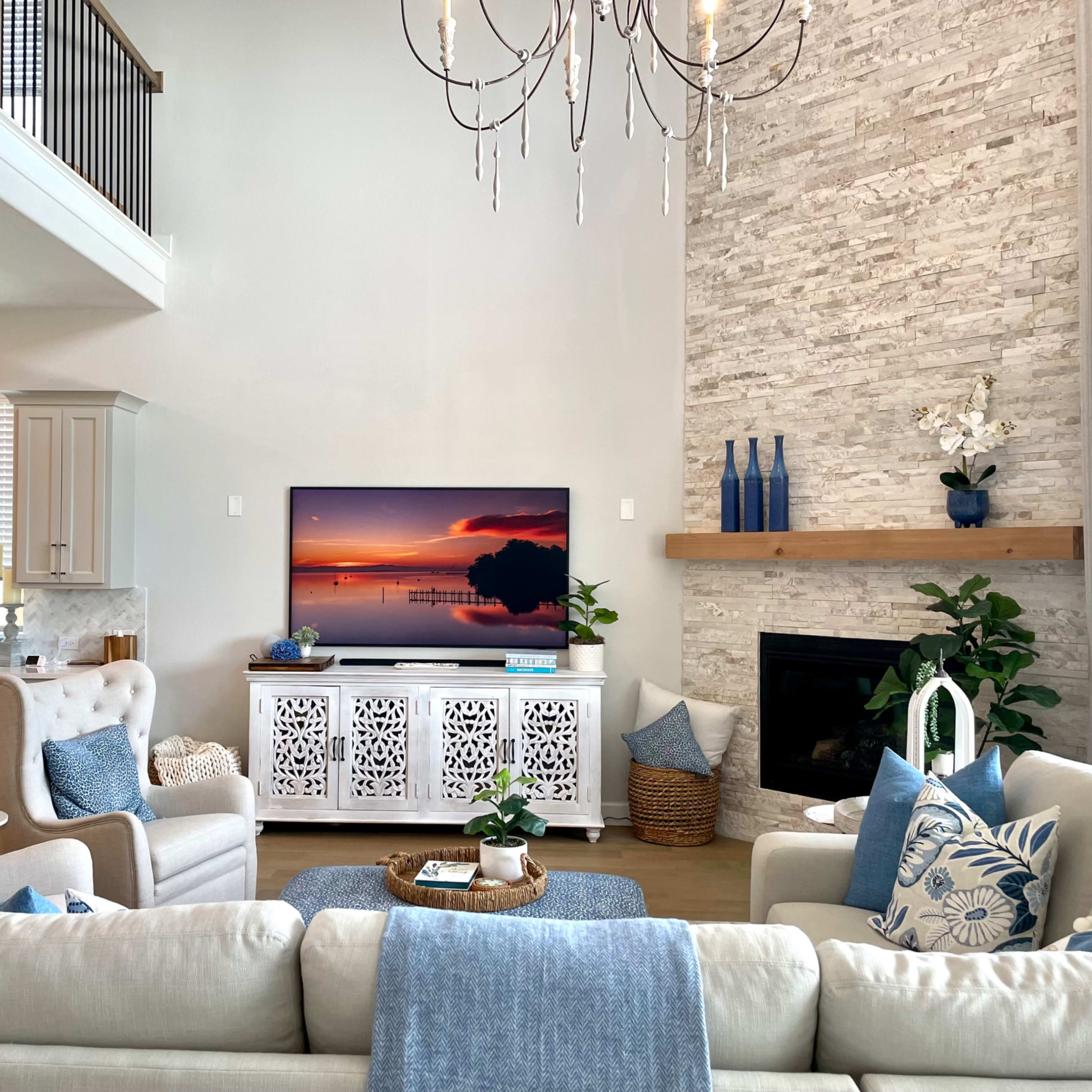 A modern living room features a large television displaying a sunset, surrounded by white furniture, a stone fireplace, and decorative plants.