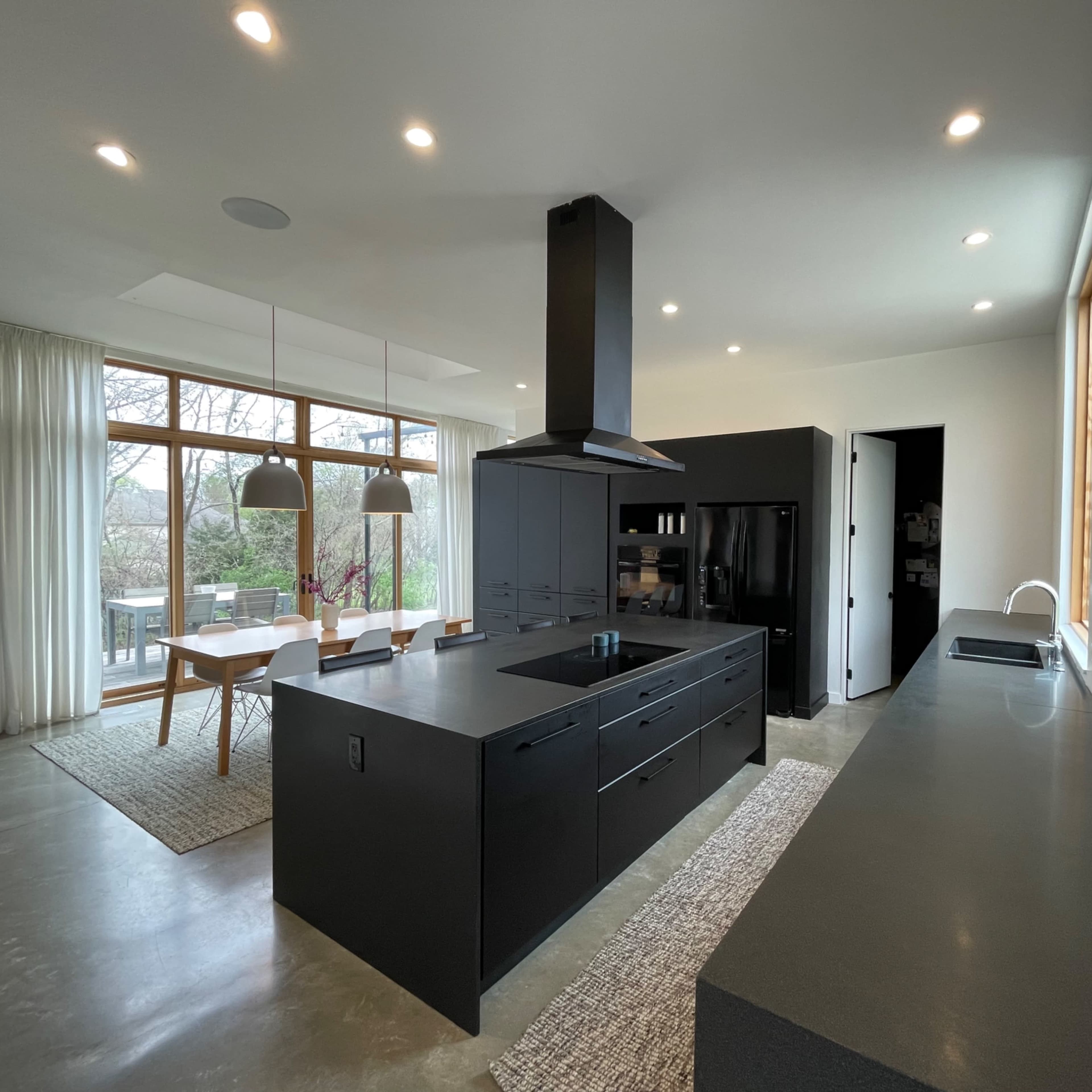 A modern kitchen features a large black island with an integrated sink, sleek cabinetry, and a ventilation hood, with natural light streaming through large windows.