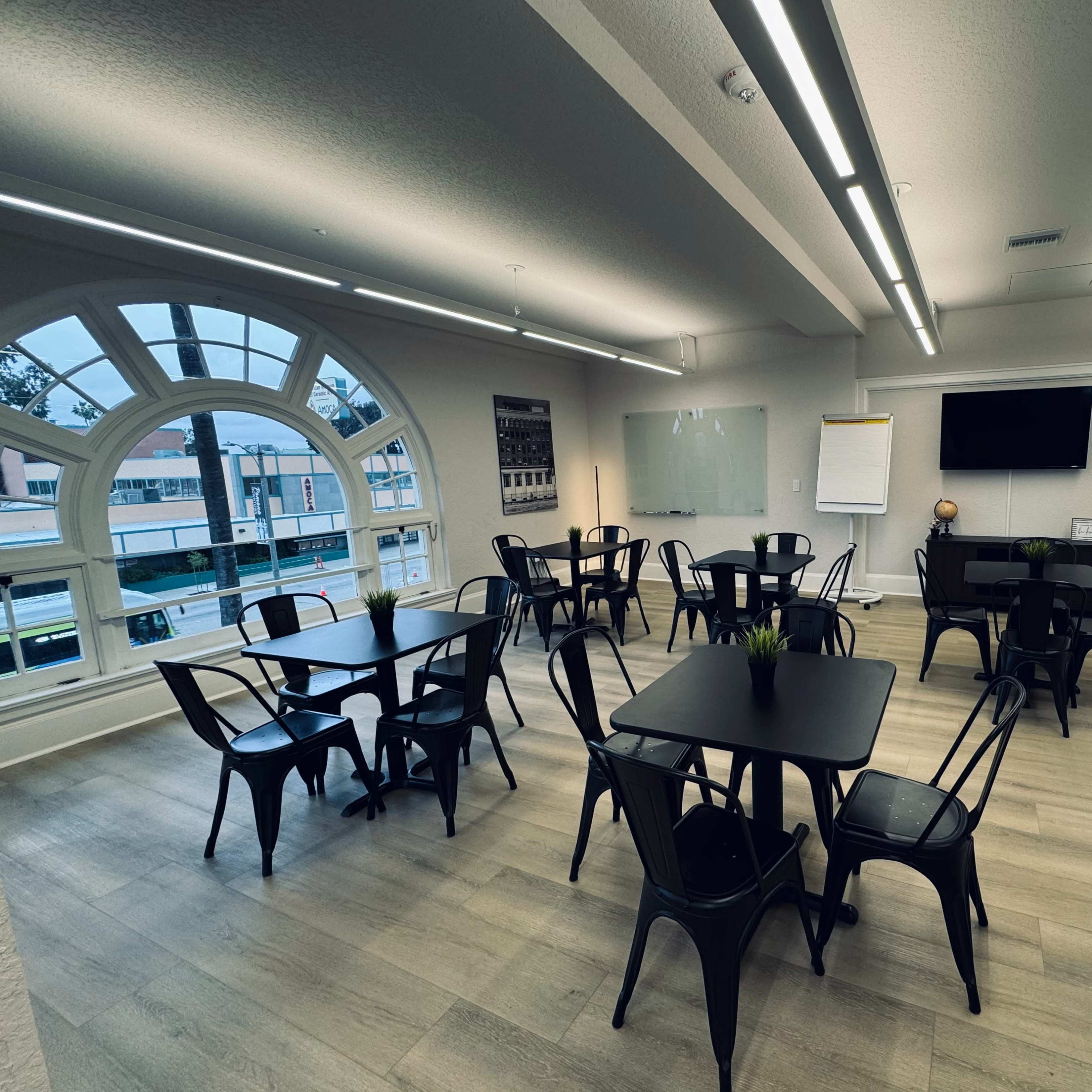 A meeting room features several black tables and chairs, large windows, and a presentation screen on the wall.