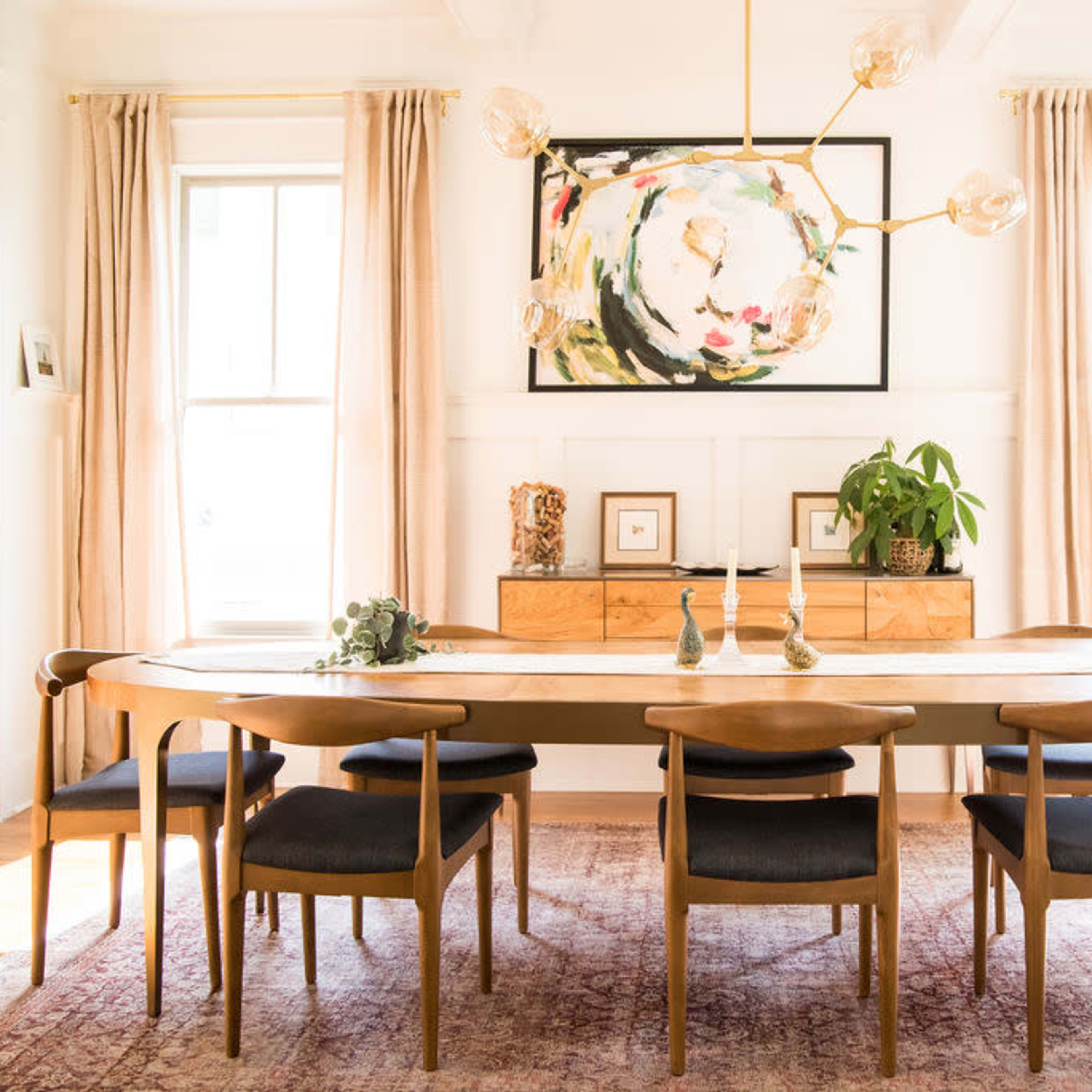 A modern dining room features a long wooden table surrounded by chairs, with large windows, artwork on the wall, and plants in the corners.