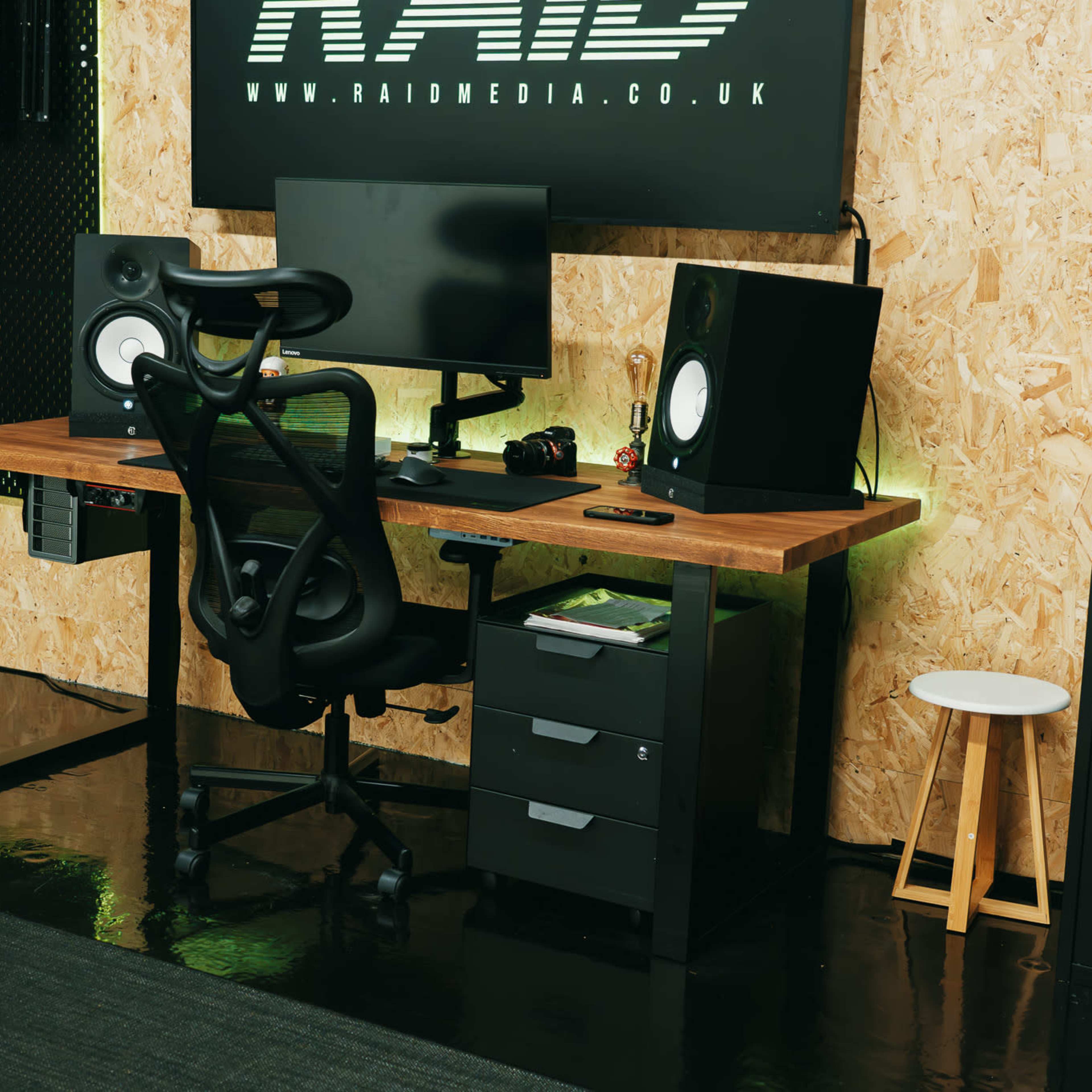 A modern desk setup features a computer monitor, speakers, an ergonomic chair, and storage drawers against a textured wooden wall.