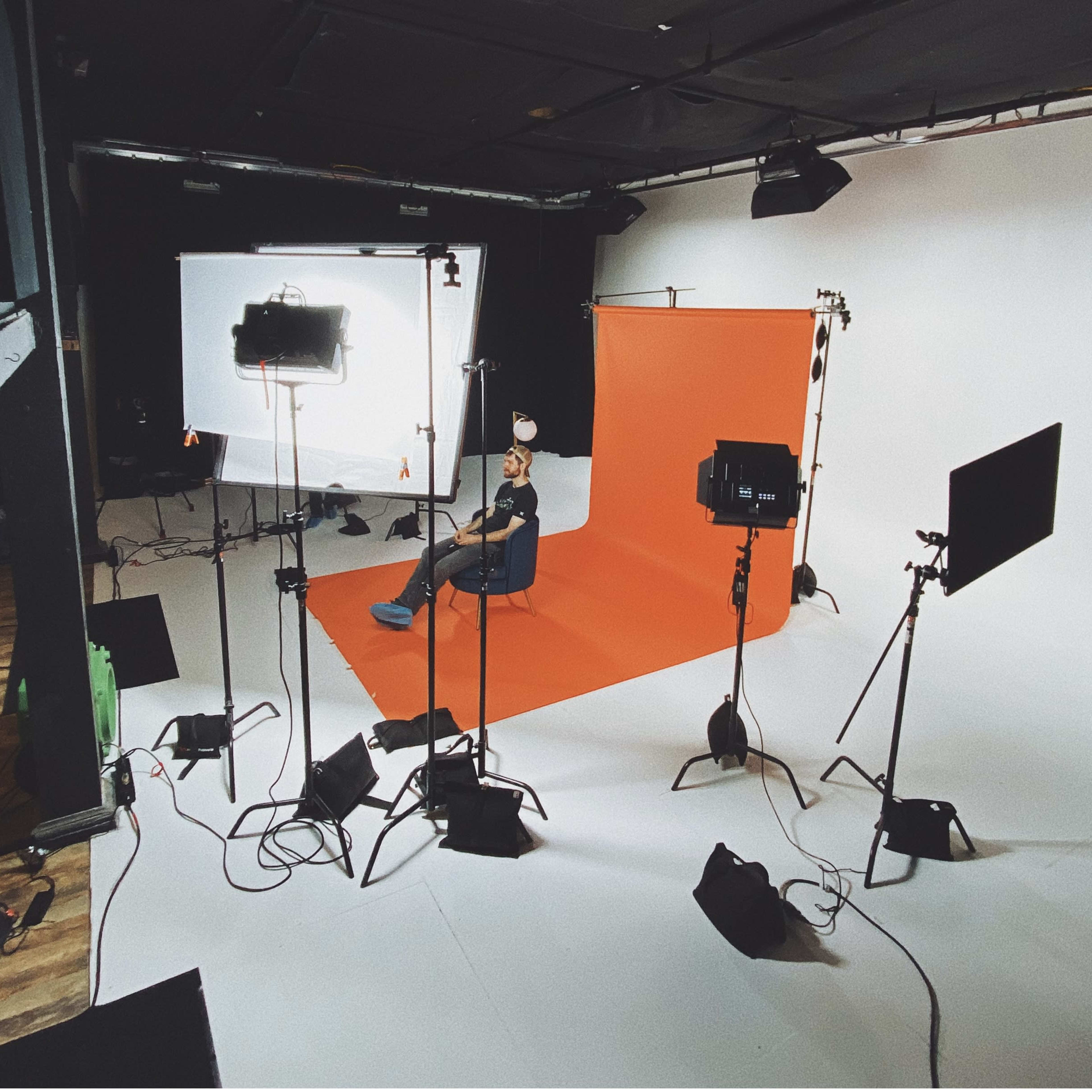 A studio setup features an orange backdrop with lighting equipment arranged around a seated individual.