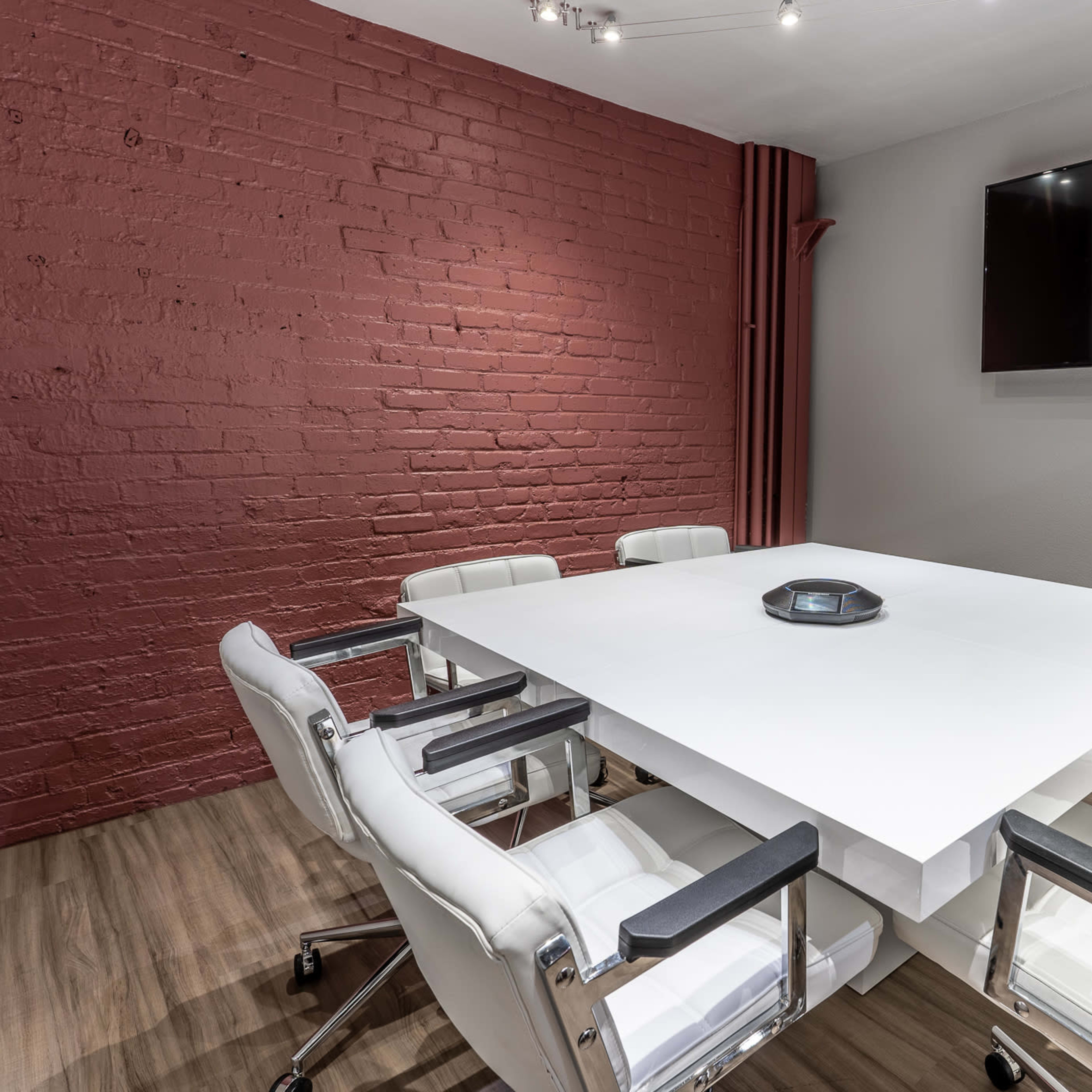 A modern conference room features a large white table surrounded by white chairs, with a red brick wall and a mounted TV.