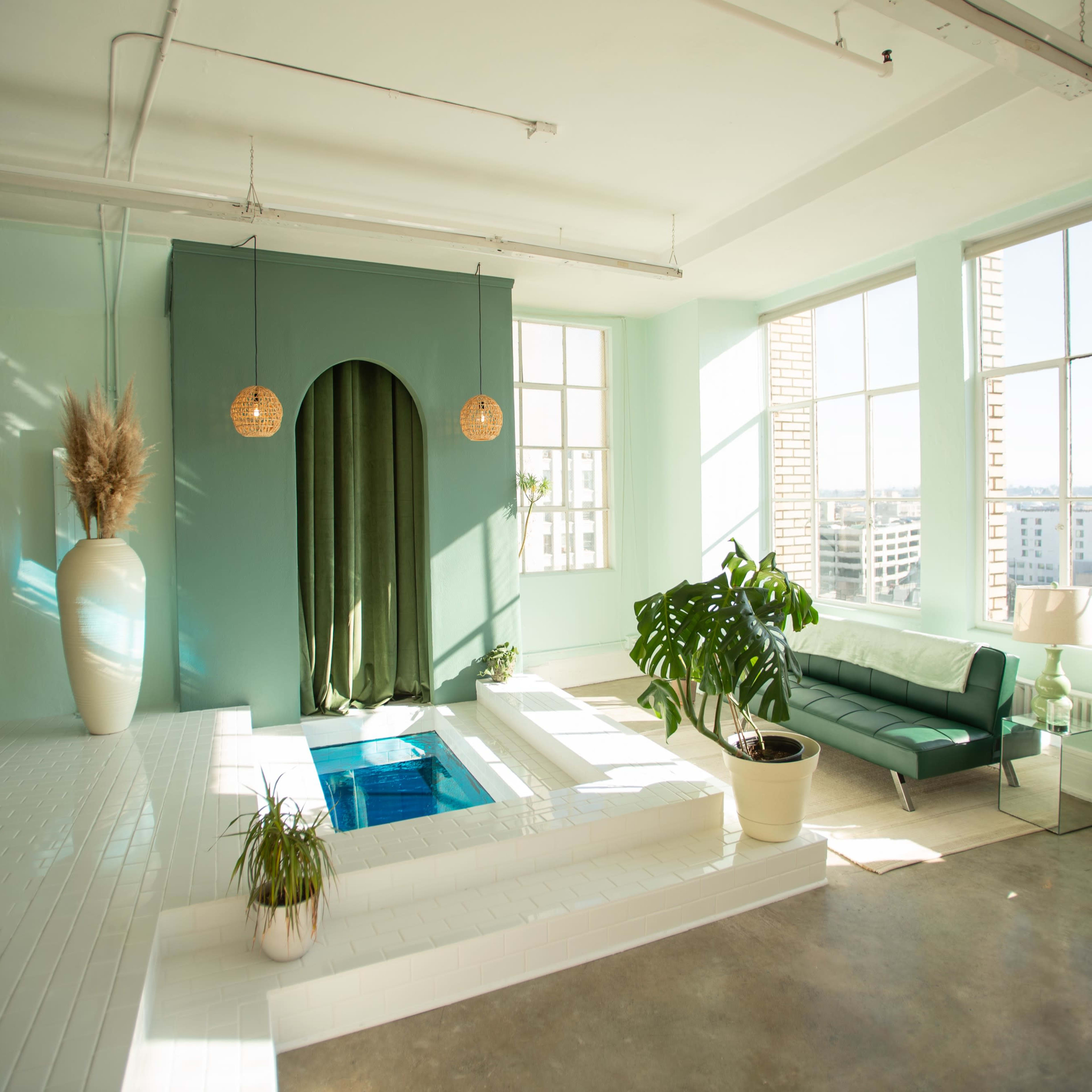 A modern interior space features a small indoor pool surrounded by plants and a turquoise wall, with large windows letting in natural light.