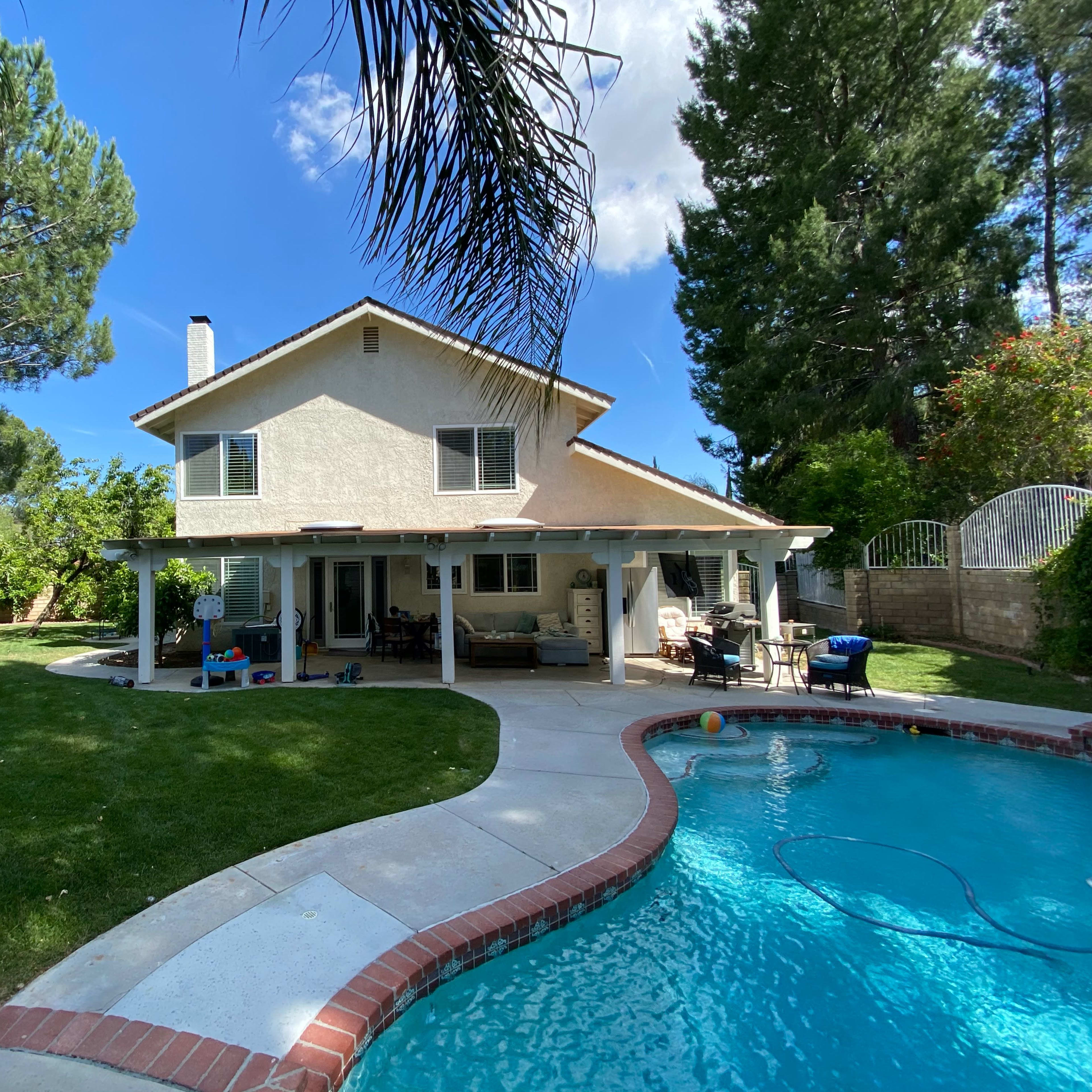 A two-story house with a pool and landscaped yard is surrounded by trees and a fence.