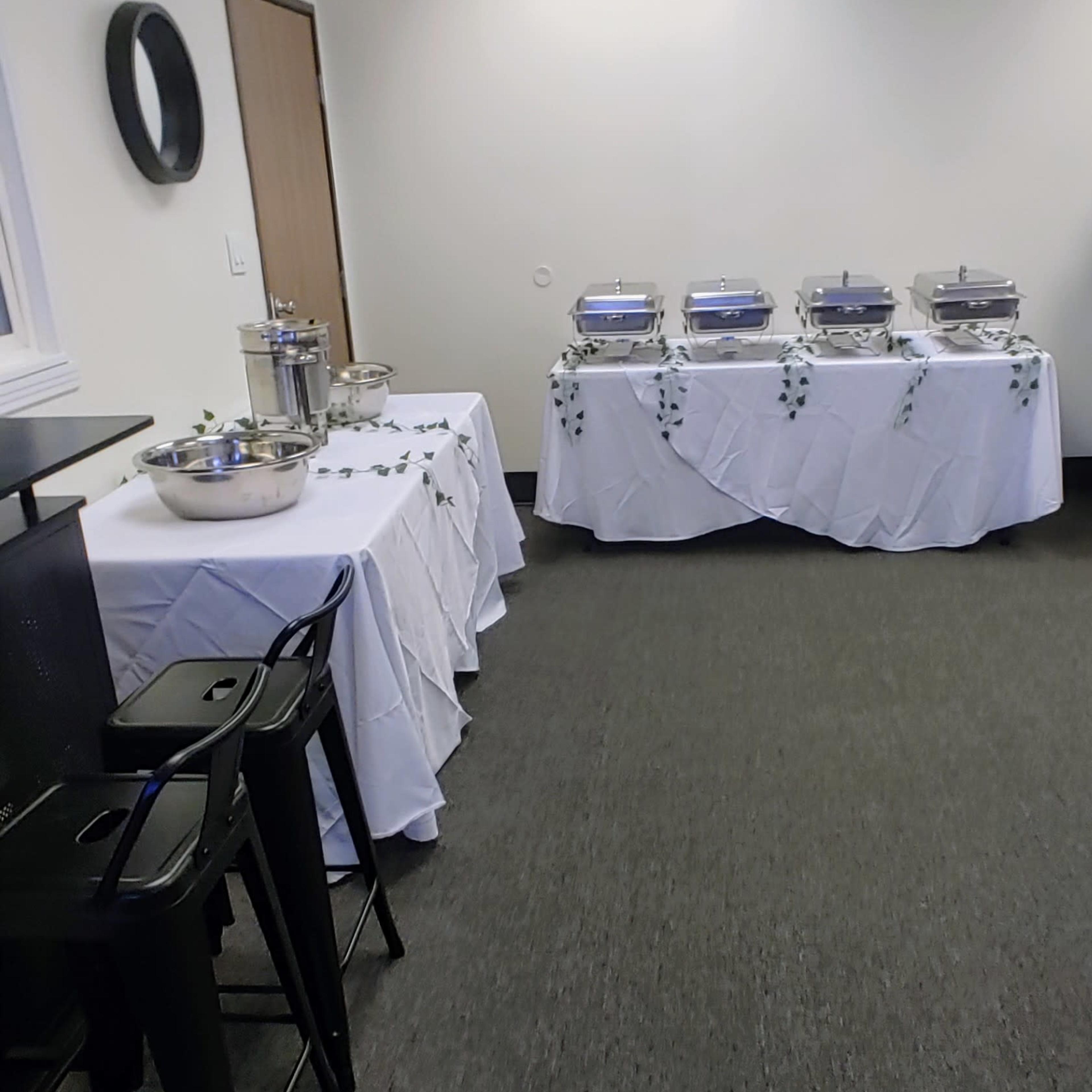 A room set up for an event, featuring two tables covered with white tablecloths, each adorned with decorative cloths and equipped with chafing dishes and bowls.