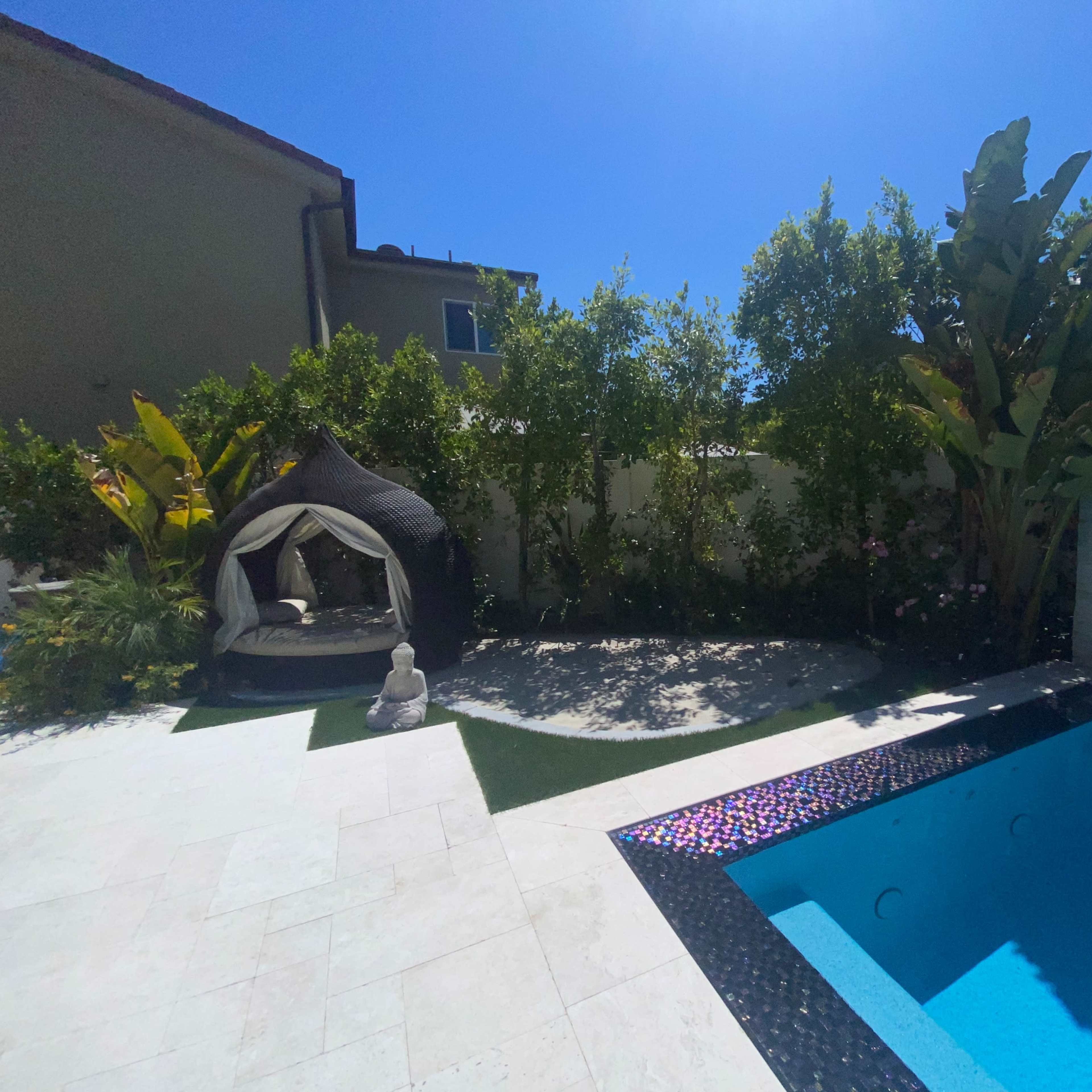 A backyard with a pool, a lounging area featuring a canopy, and lush greenery alongside a stone path.