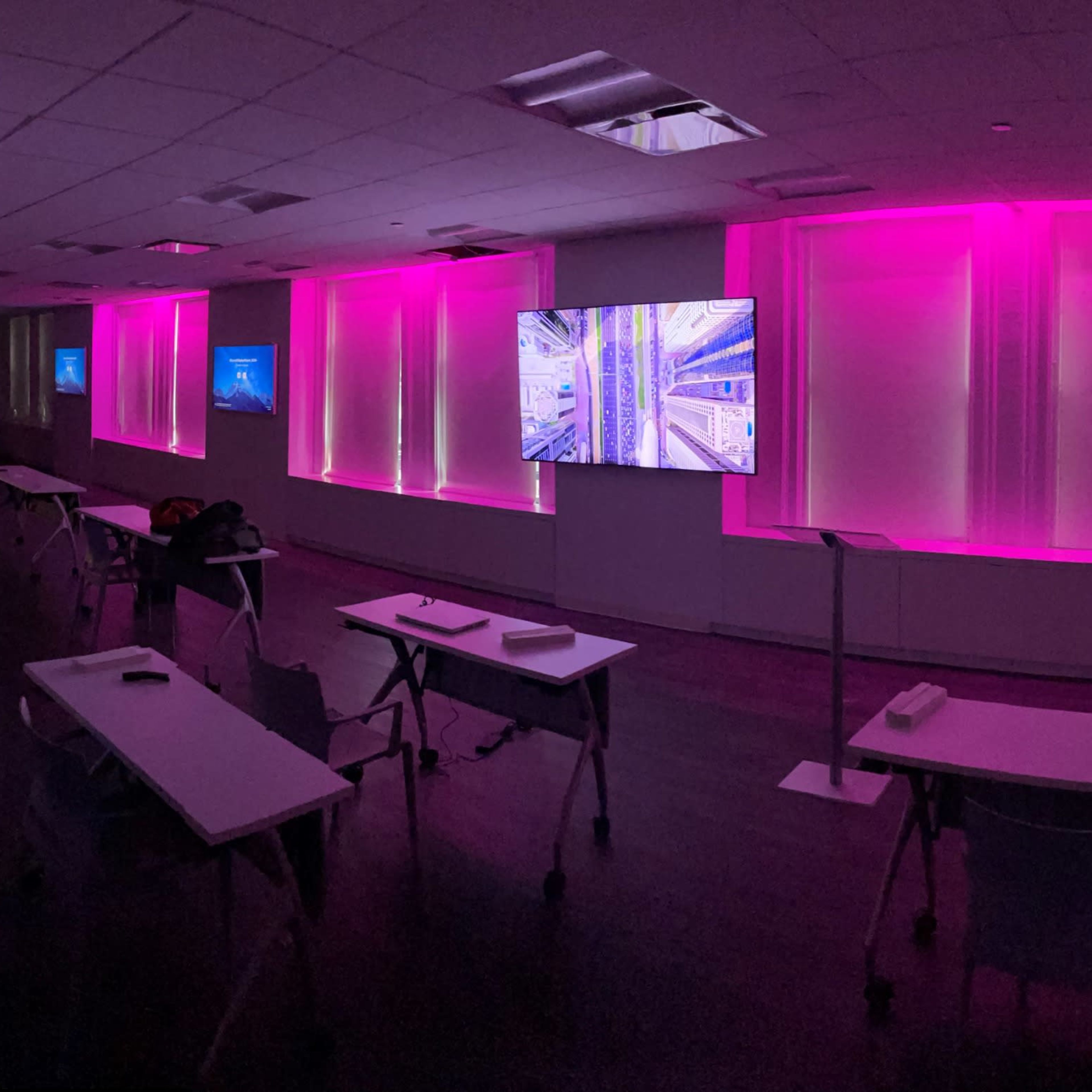 A modern, dimly lit room with multiple screens displaying abstract visuals, surrounded by pink ambient lighting and arranged tables facing the displays.