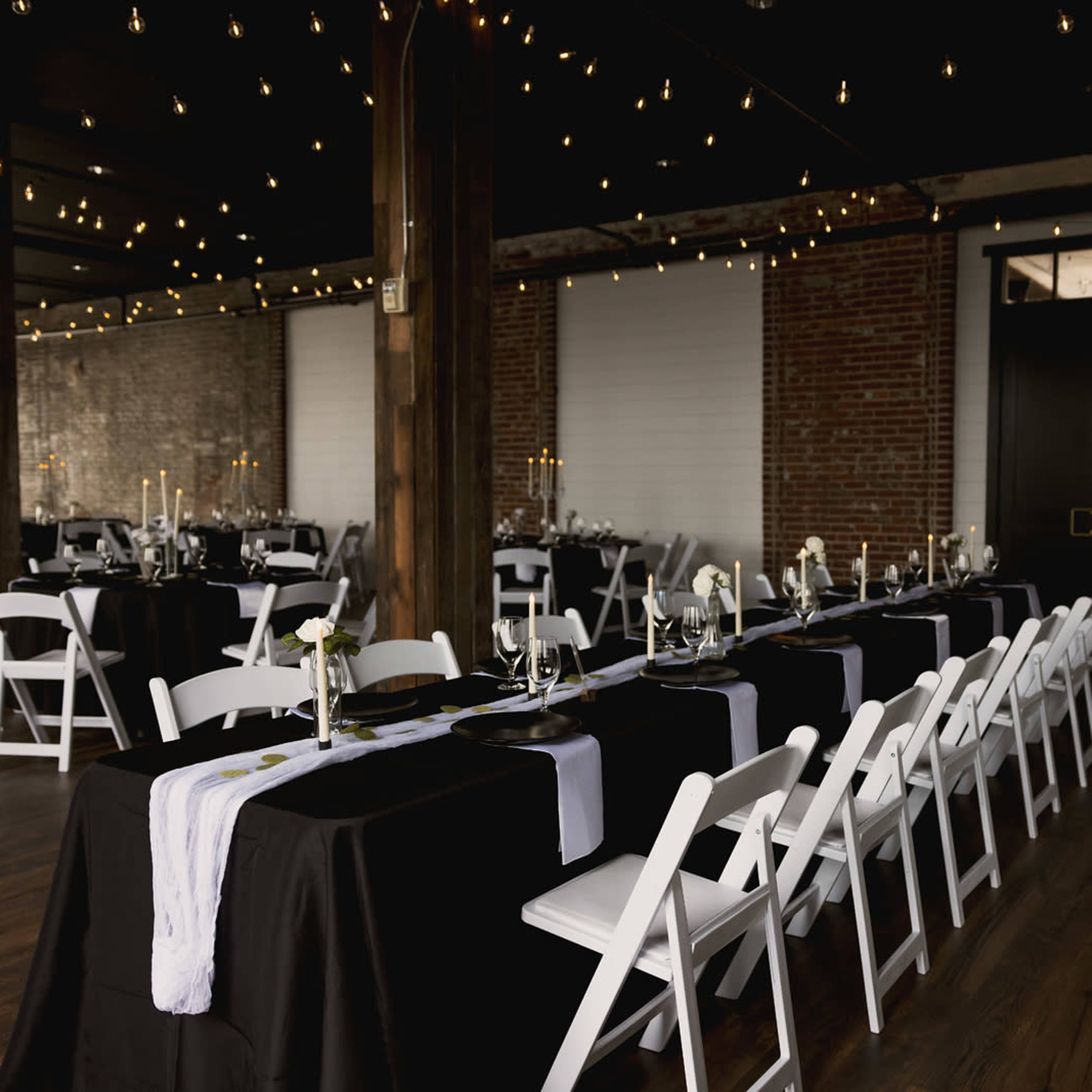 A decorated event space with long tables set with black and white tablecloths, white chairs, and ambient lighting from hanging string lights.