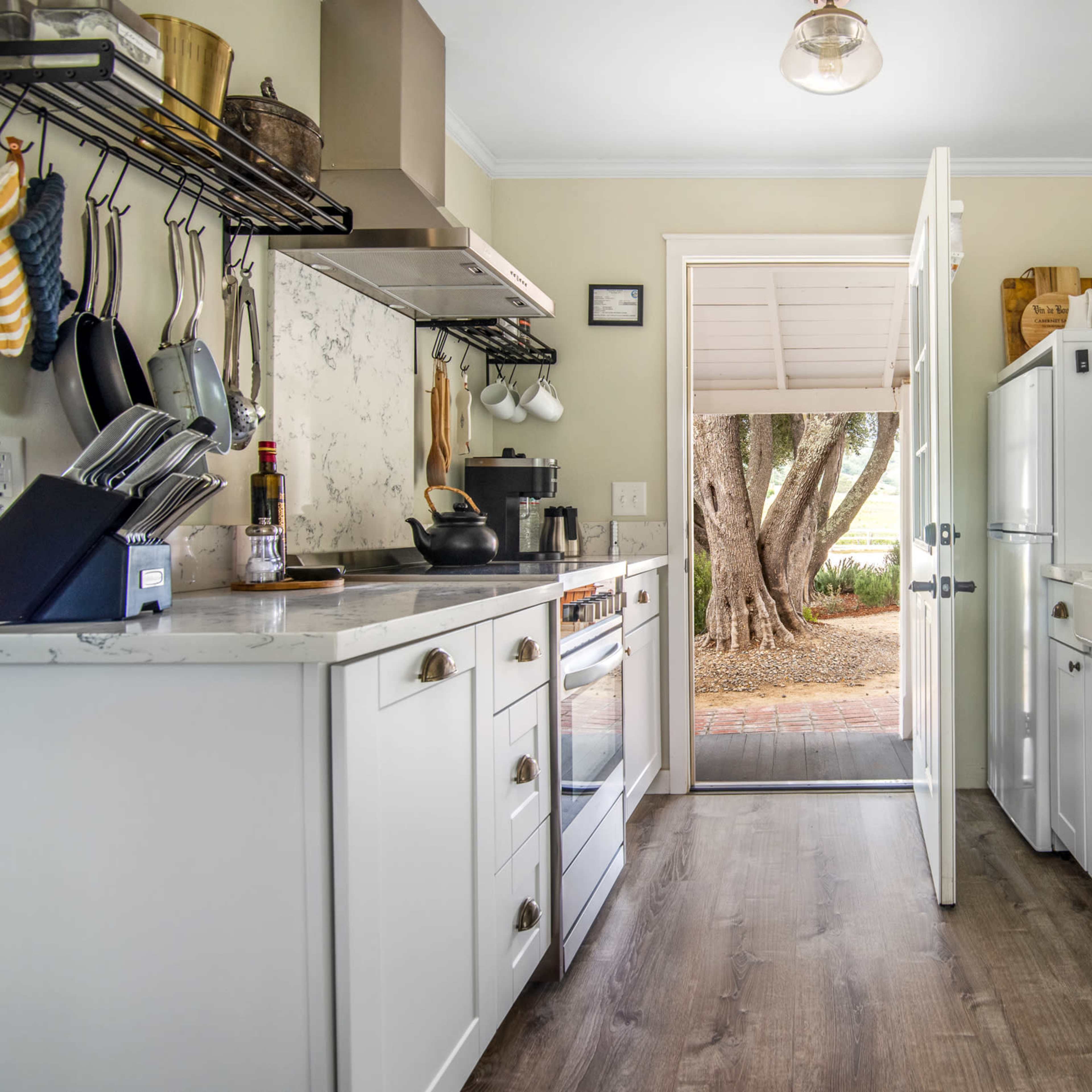 A kitchen with white cabinetry opens to an outdoor area through a door, featuring a side-by-side refrigerator and shelves with glassware.