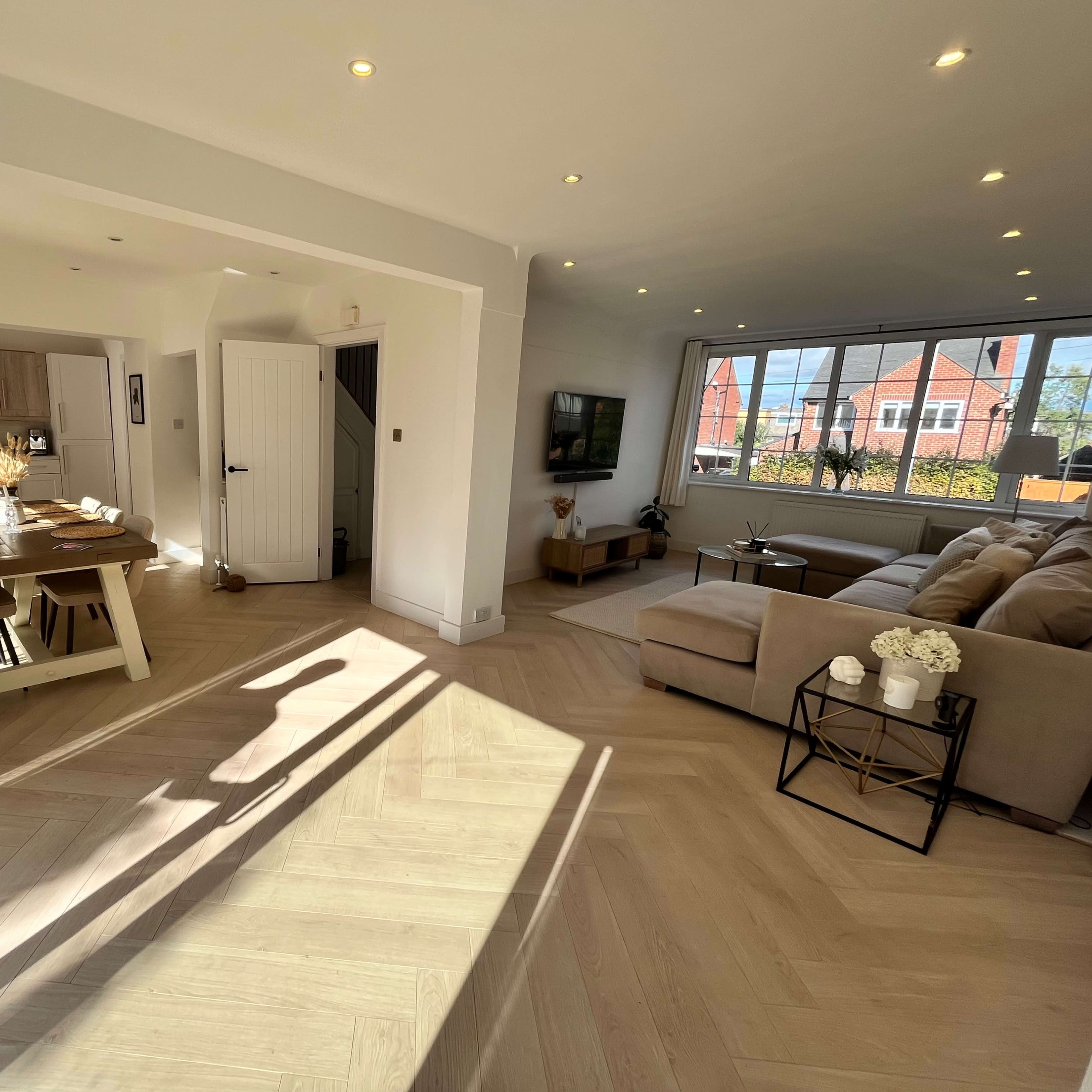 A modern open-plan living area with a dining table, a large sectional sofa, and large windows allowing natural light to fill the space.