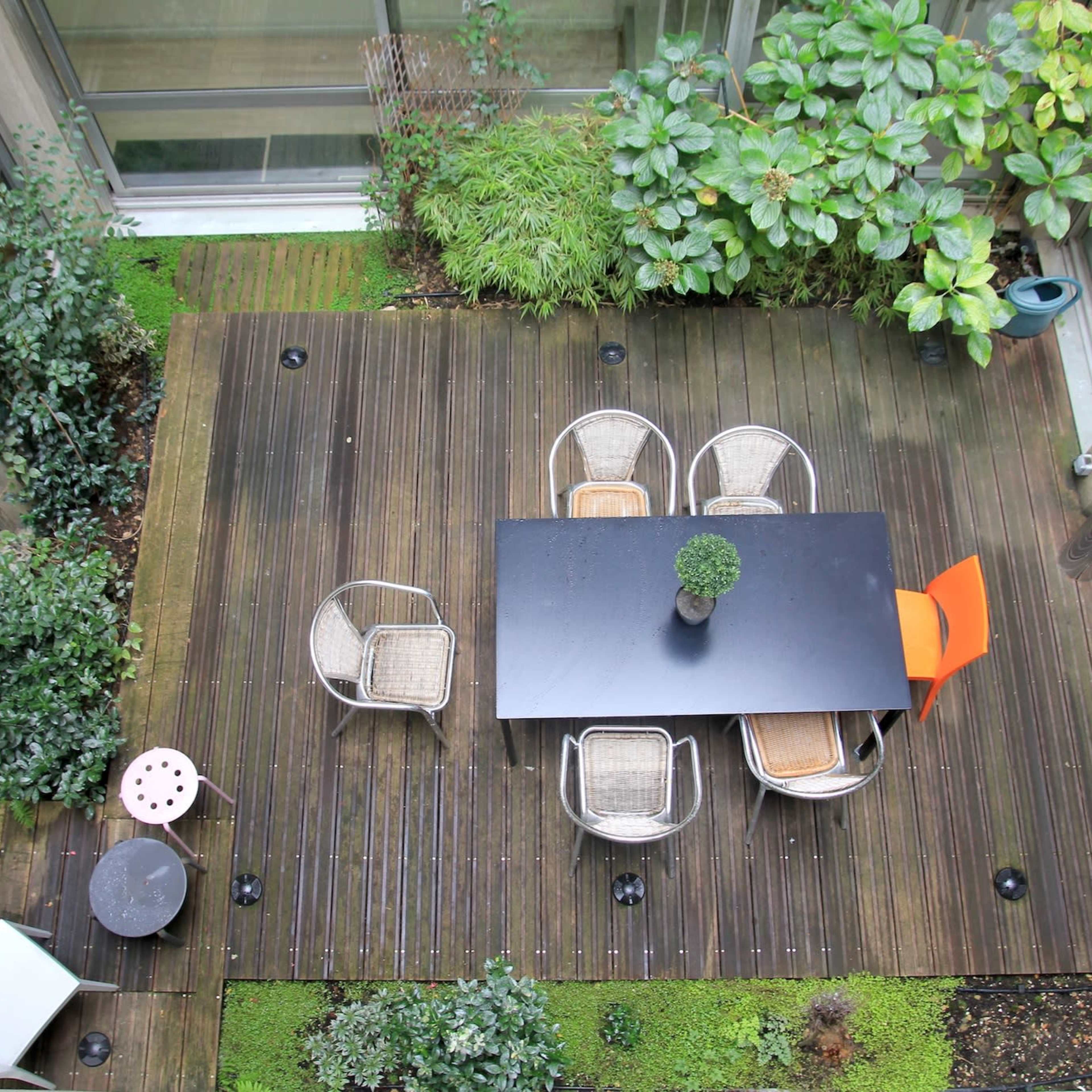 A wooden deck with a black table and six chairs, surrounded by greenery and planters.