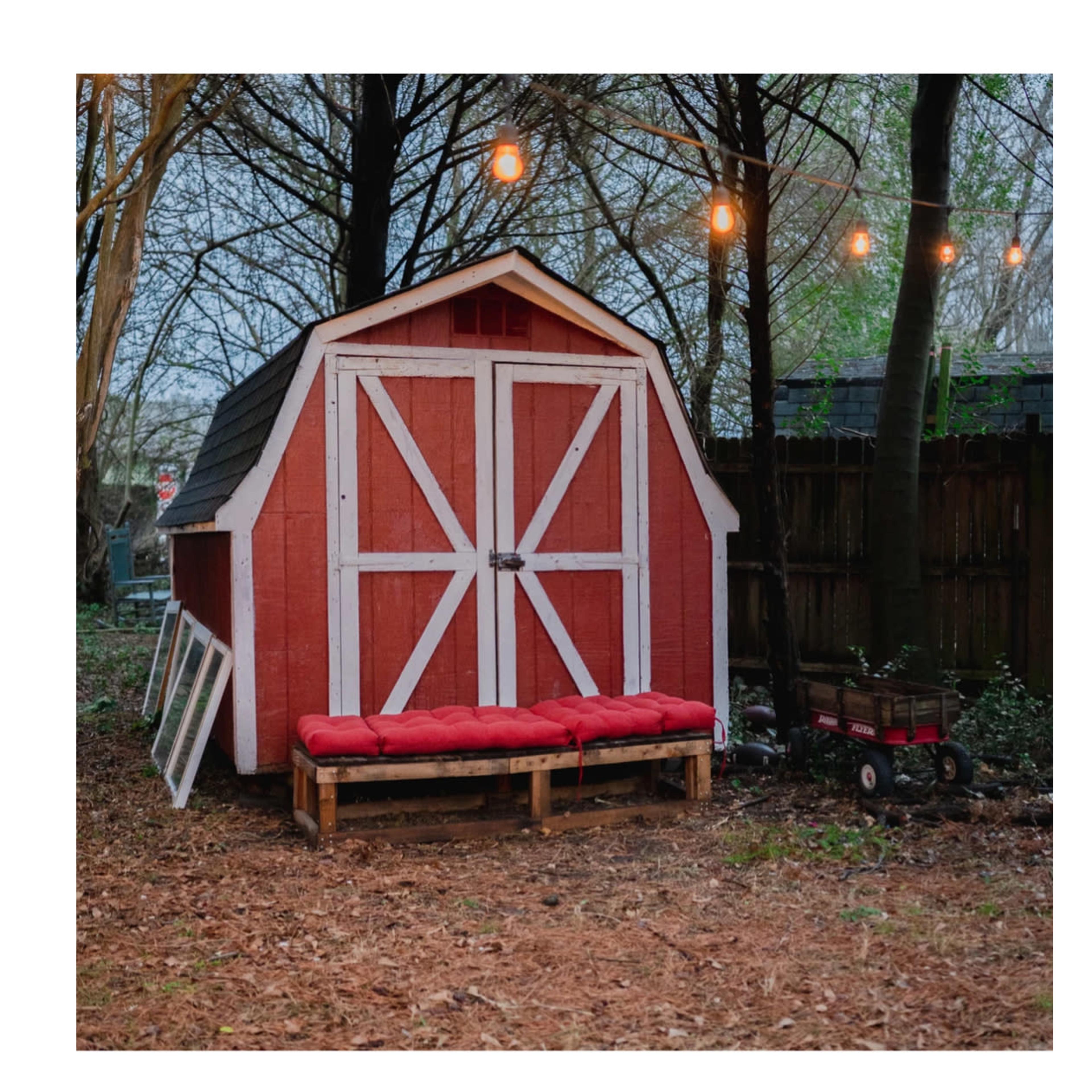 A red barn-style shed with white trim and a wooden bench sits in a wooded area illuminated by string lights.