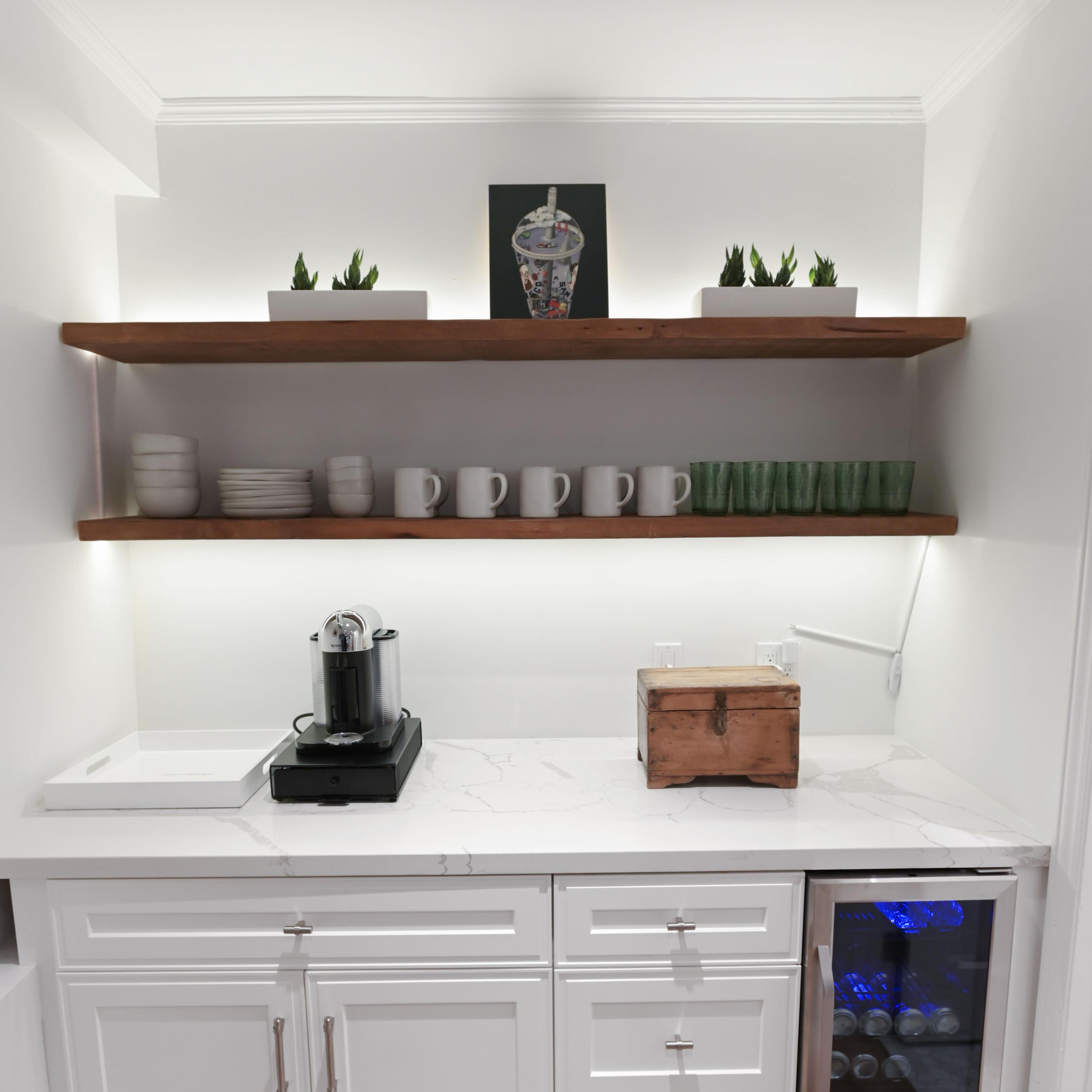 A modern kitchen area features a countertop with a coffee machine, mugs, a wooden box, and a mini refrigerator below a wall-mounted shelf with plants and dishware.