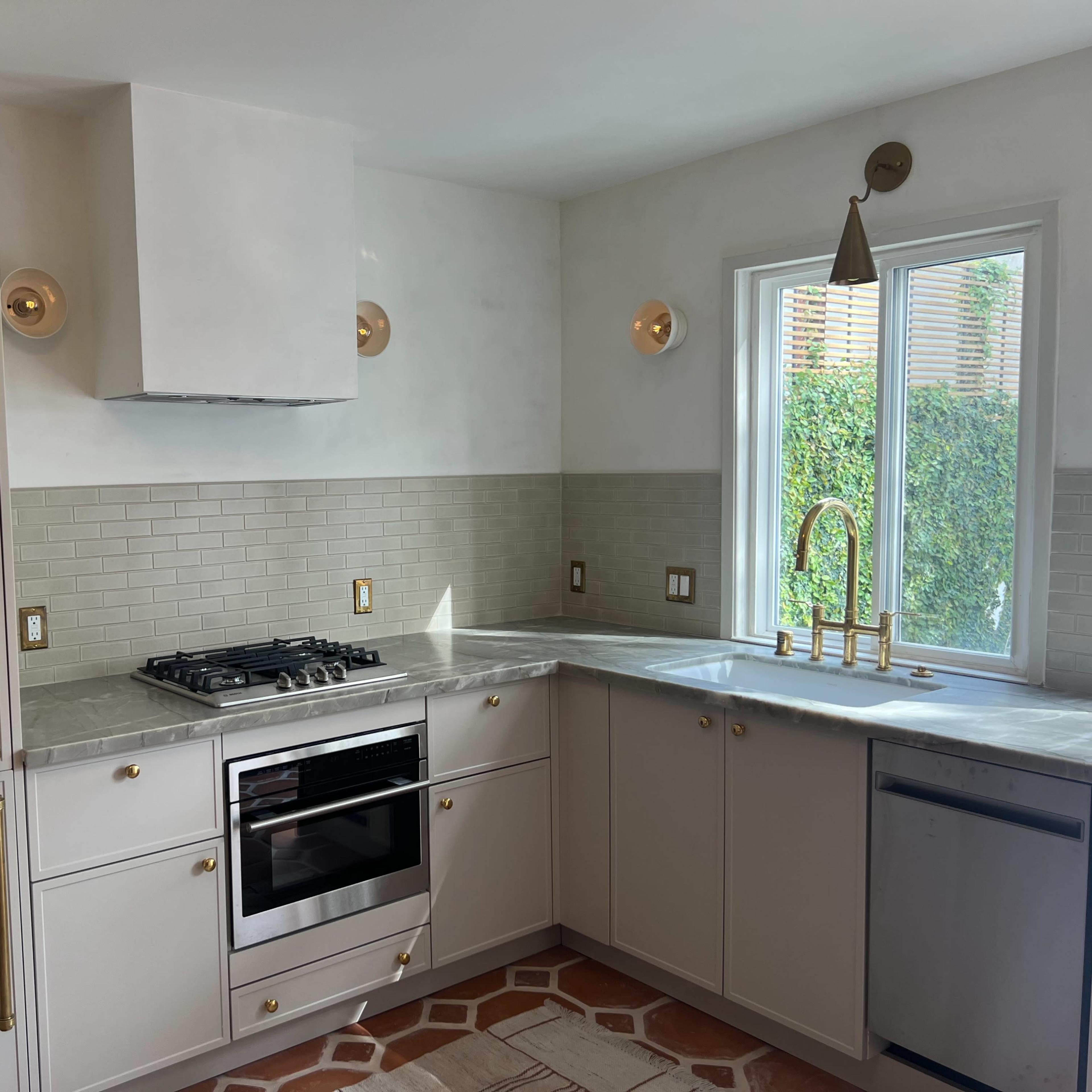 A modern kitchen with a gray countertop, stainless steel appliances, and gold fixtures, featuring light-colored cabinetry and natural light coming through a window.