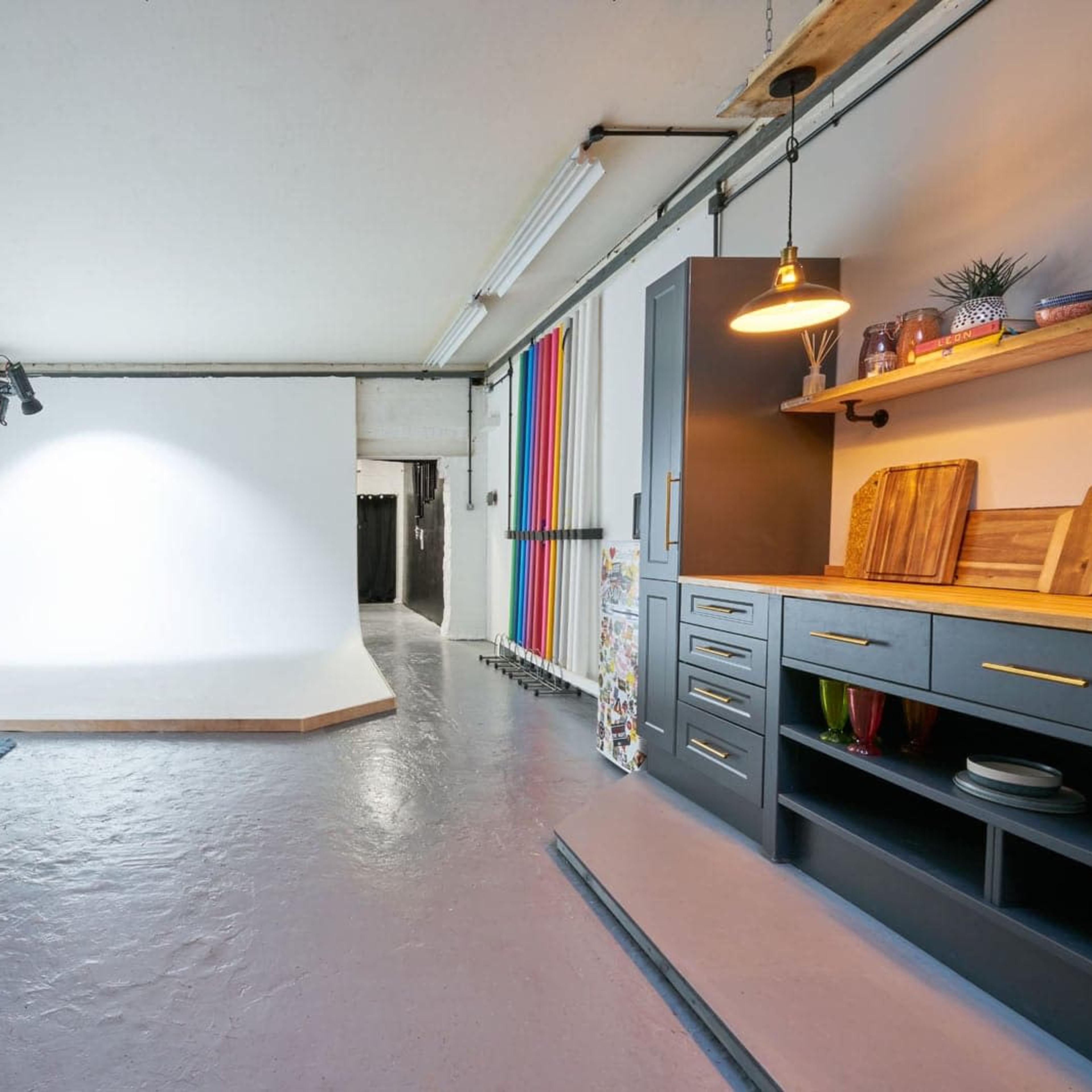 A spacious studio with a white backdrop, a yellow couch, a wooden coffee table, and a well-organized kitchen area featuring shelves and cabinets.