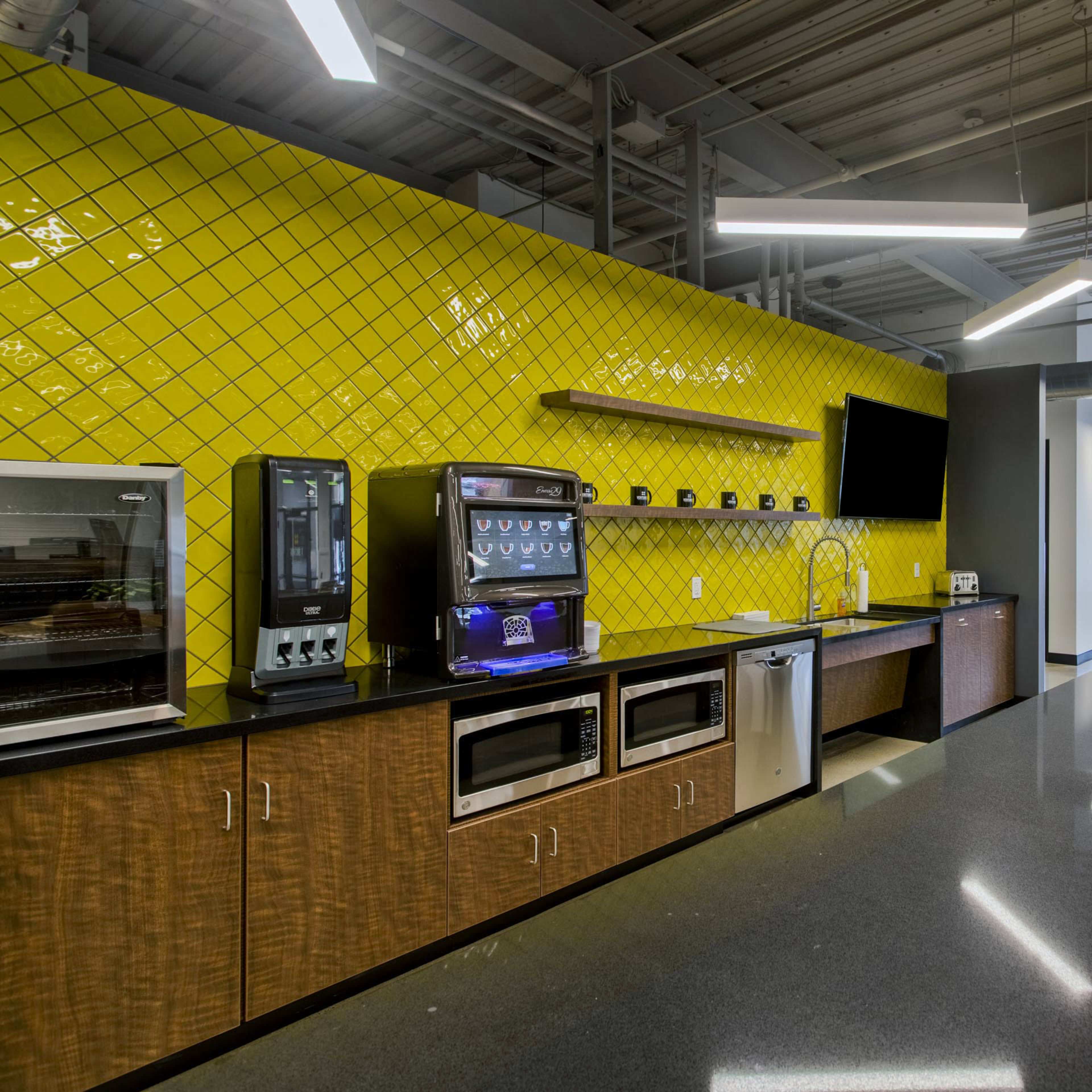 A modern kitchen area with sleek countertops, a yellow tiled wall, vending machines, and seating space.