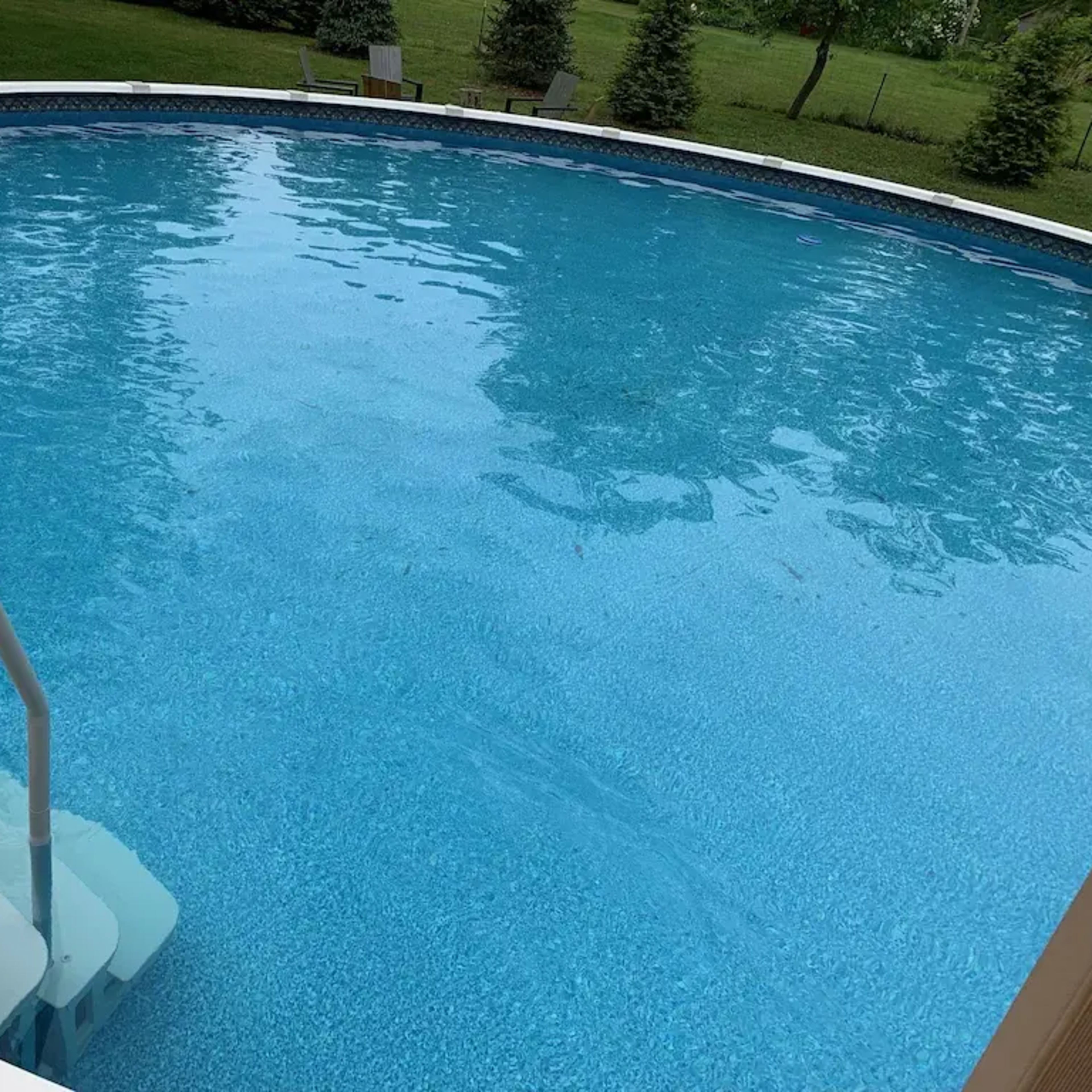 A round swimming pool with blue water and a set of white stairs leading into it, surrounded by a grassy area and trees in the background.