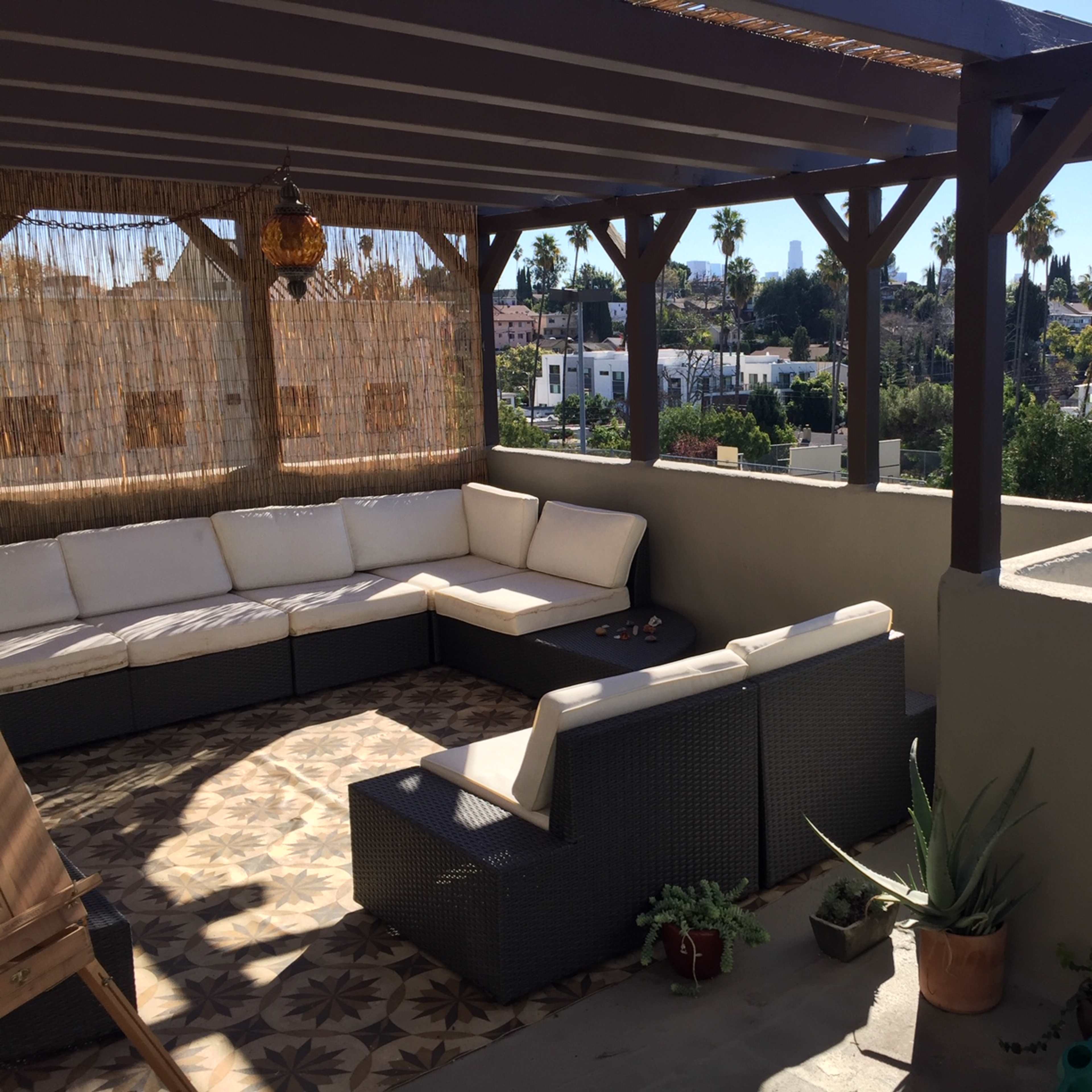 A covered outdoor seating area with a sectional sofa and potted plants overlooks a neighborhood with palm trees in the background.