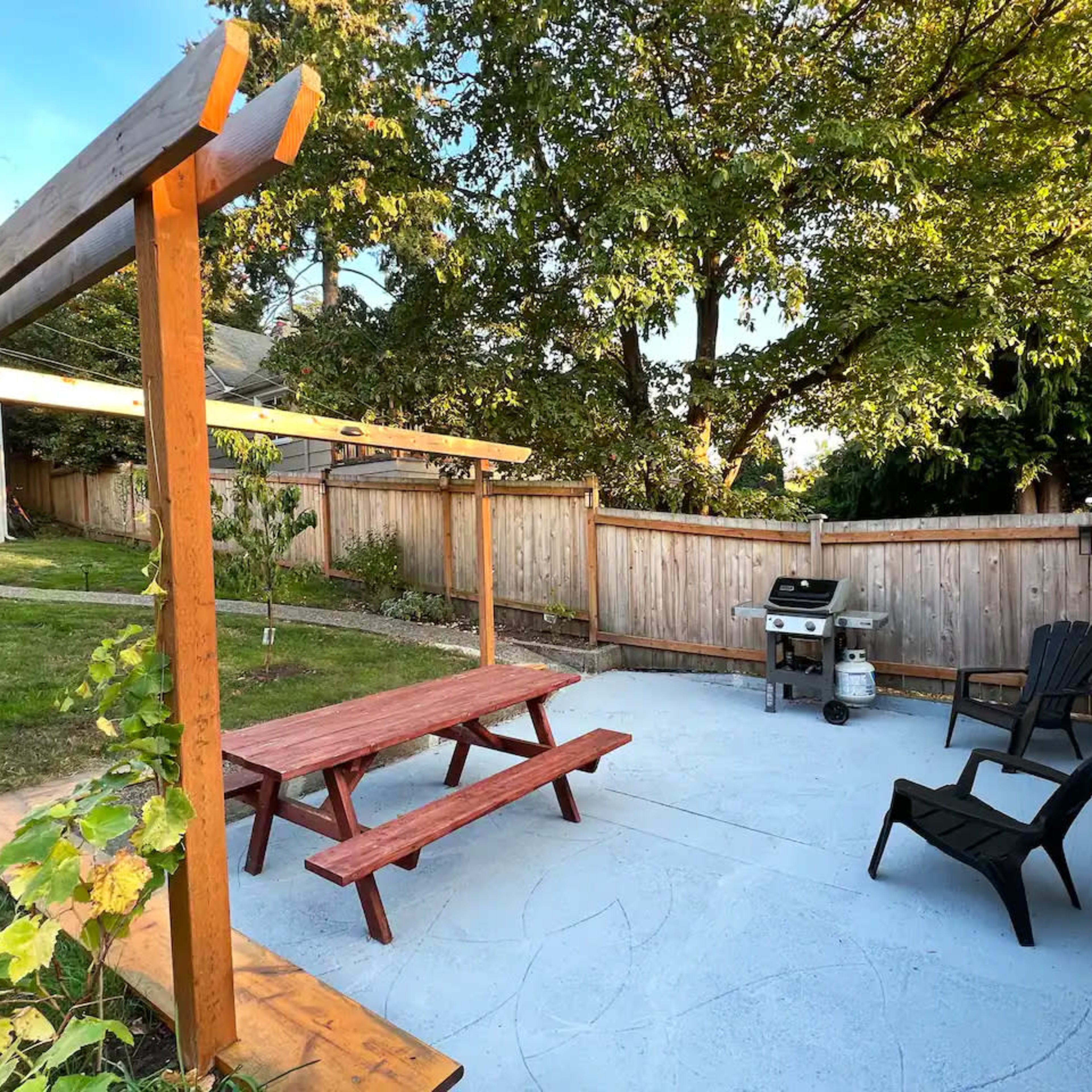 A backyard patio with a picnic table, a grill, and black Adirondack chairs surrounded by trees and a fence.