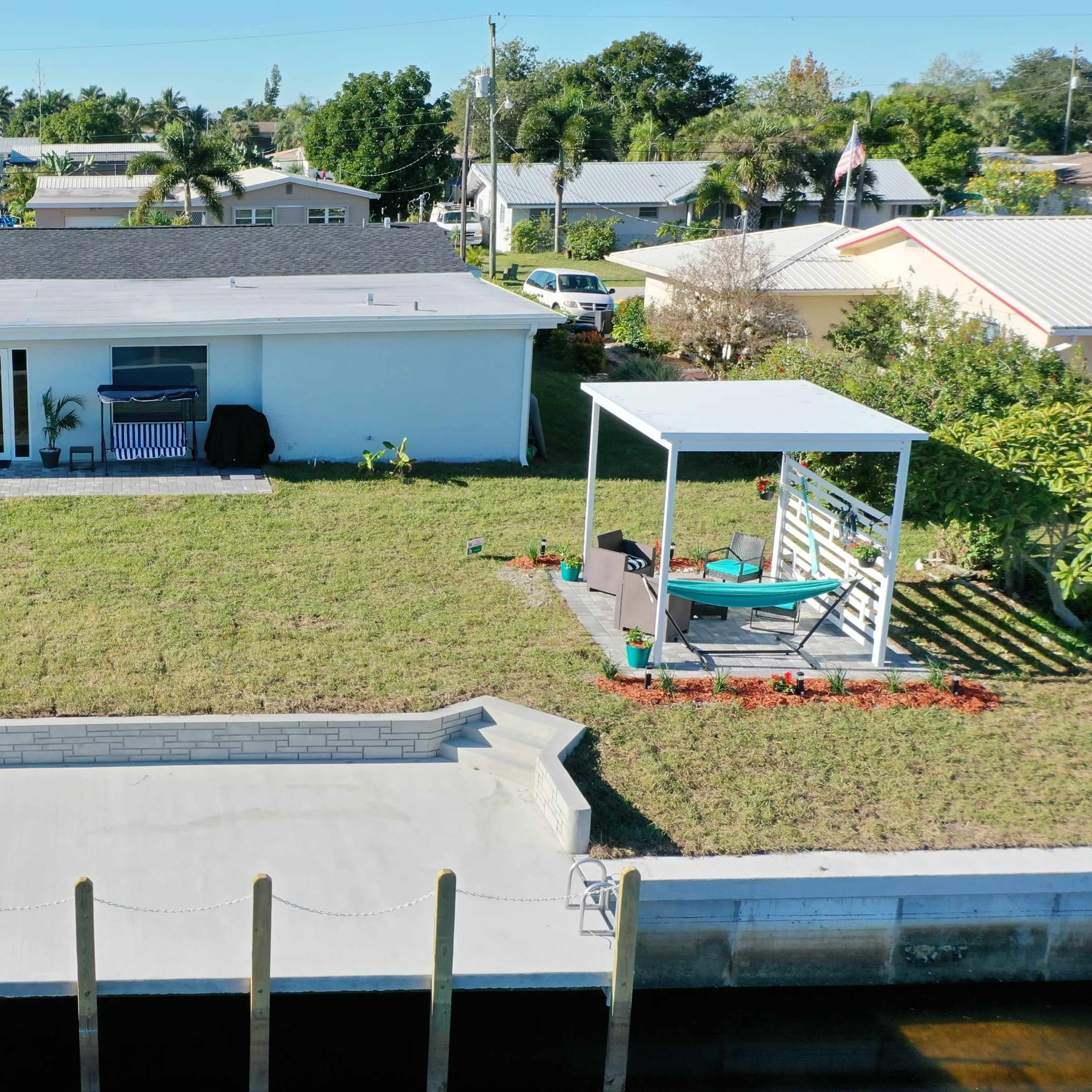 A single-story house with a back patio and a dock leading to a waterway, surrounded by greenery and residential homes.