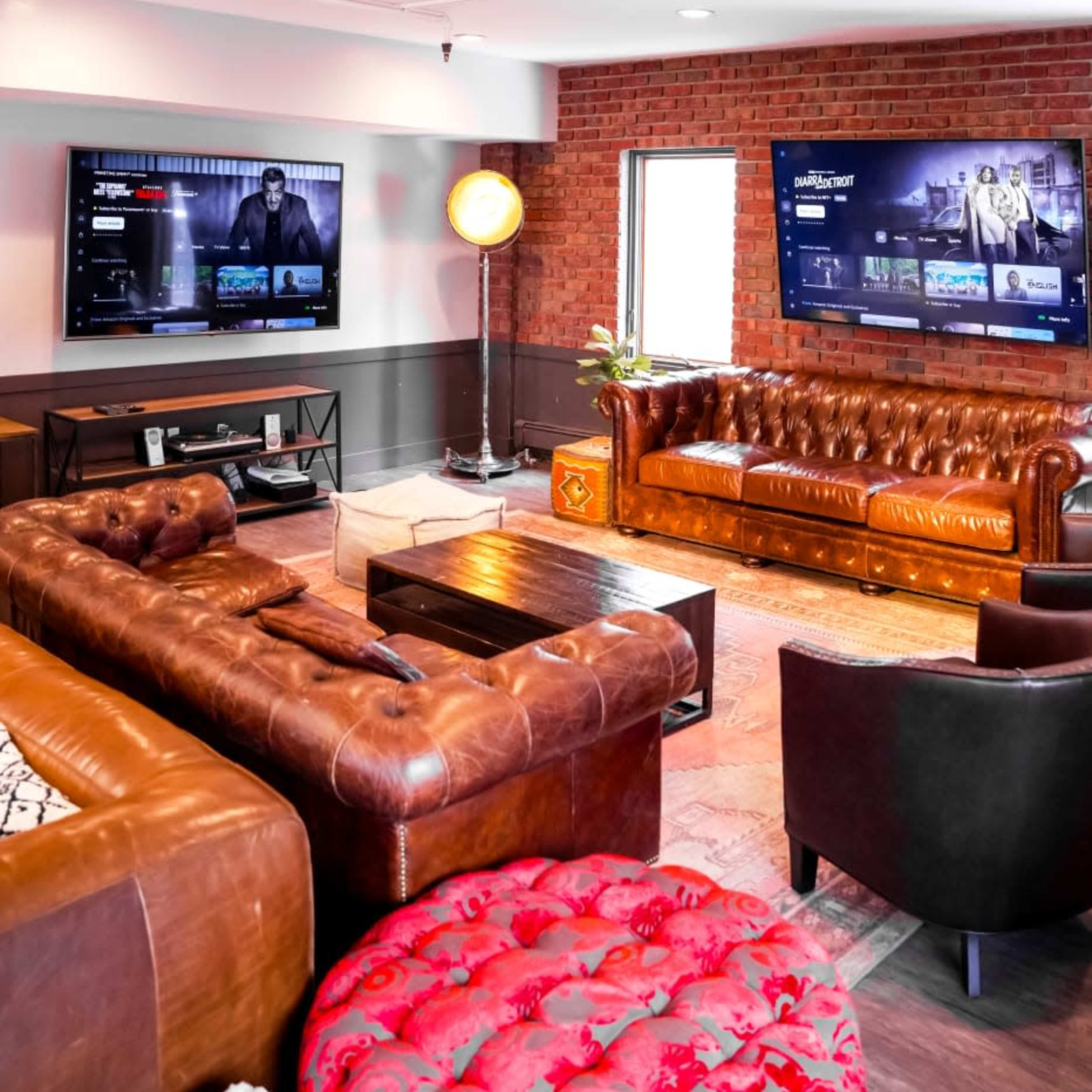 A stylish living room with two large screens mounted on a brick wall, a brown leather sofa, and a red upholstered chair.