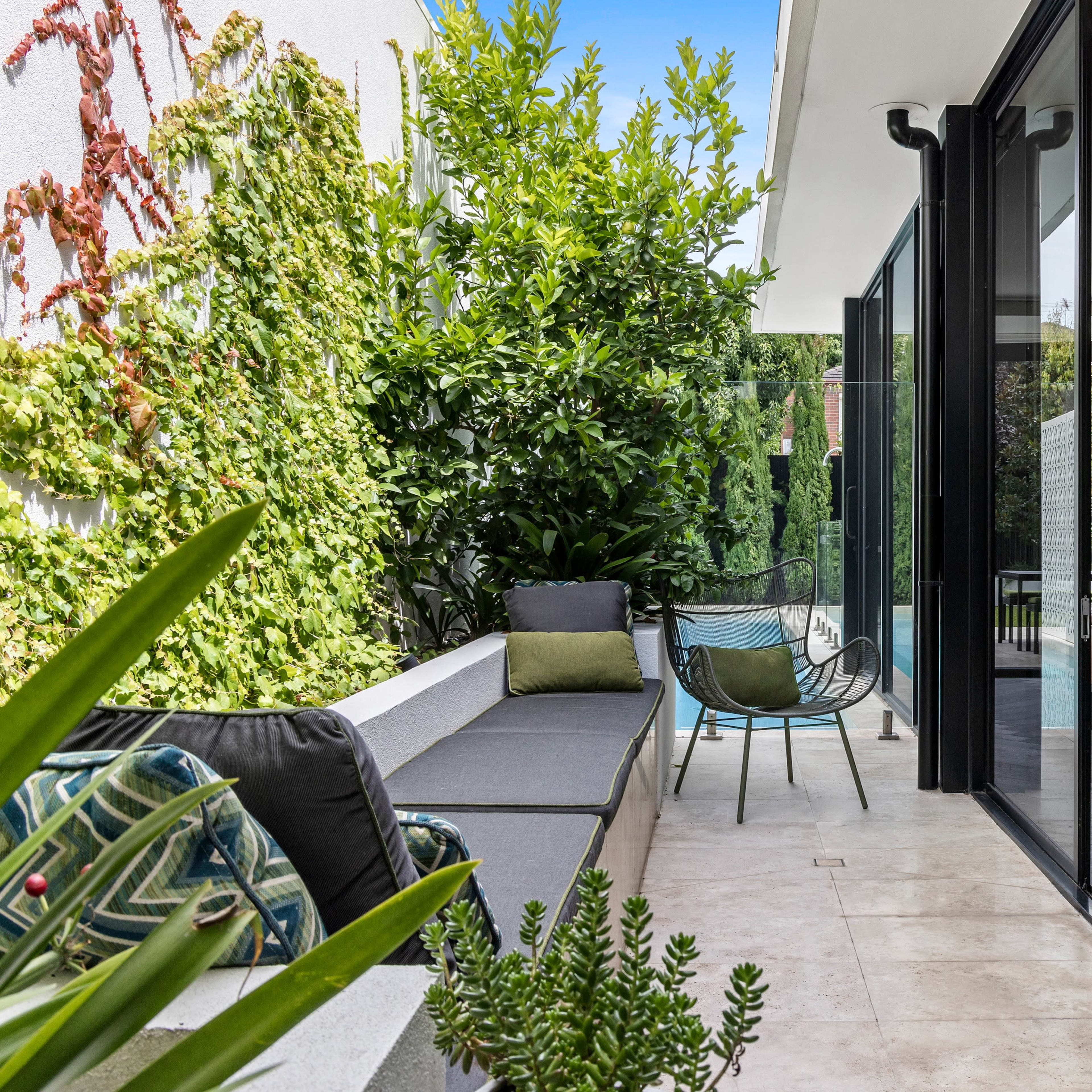 A modern patio features comfortable seating, lush greenery, and large glass doors leading to an interior space.