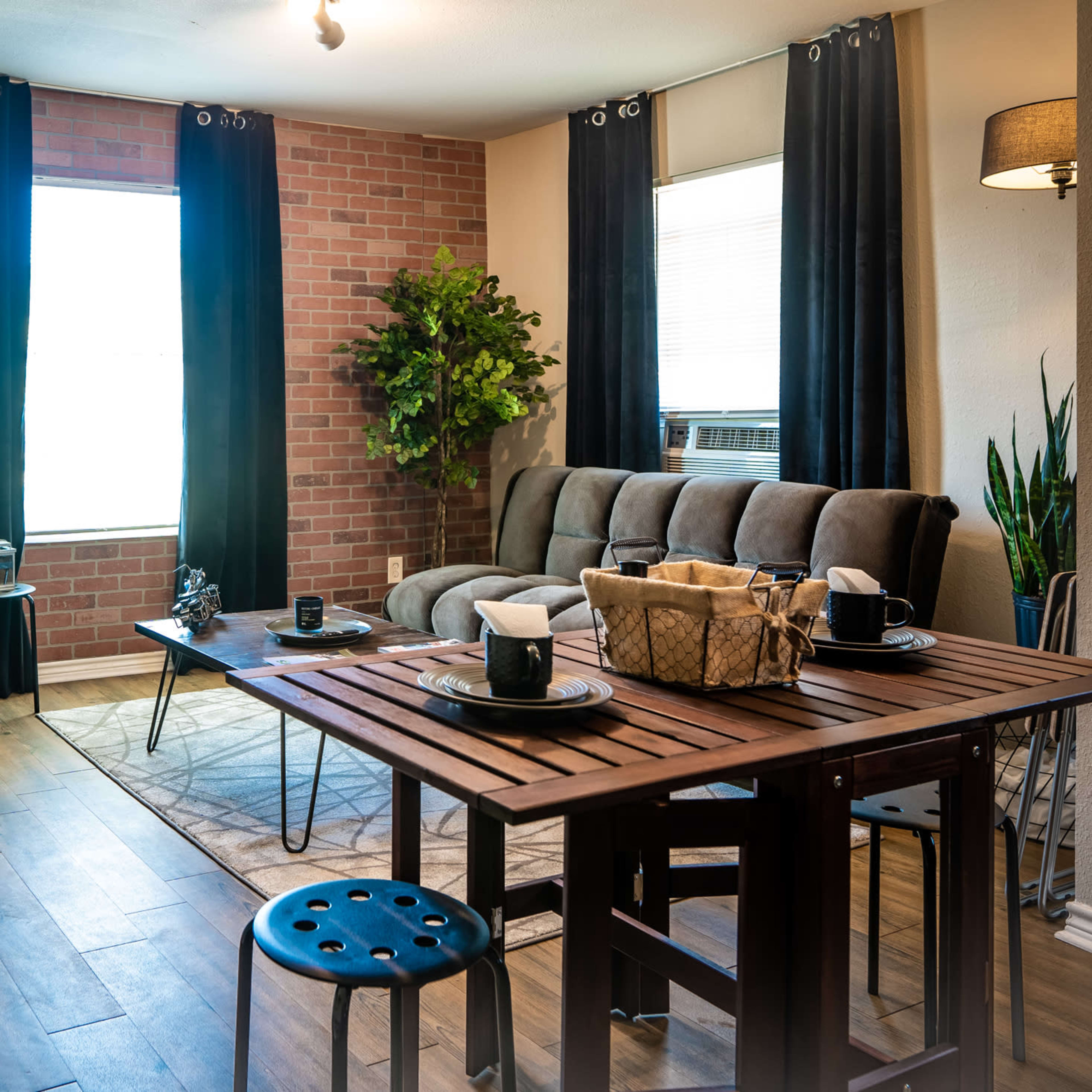 The living space features a cozy seating area with a gray couch, a wooden dining table set for four, and plants arranged against brick and windowed walls.