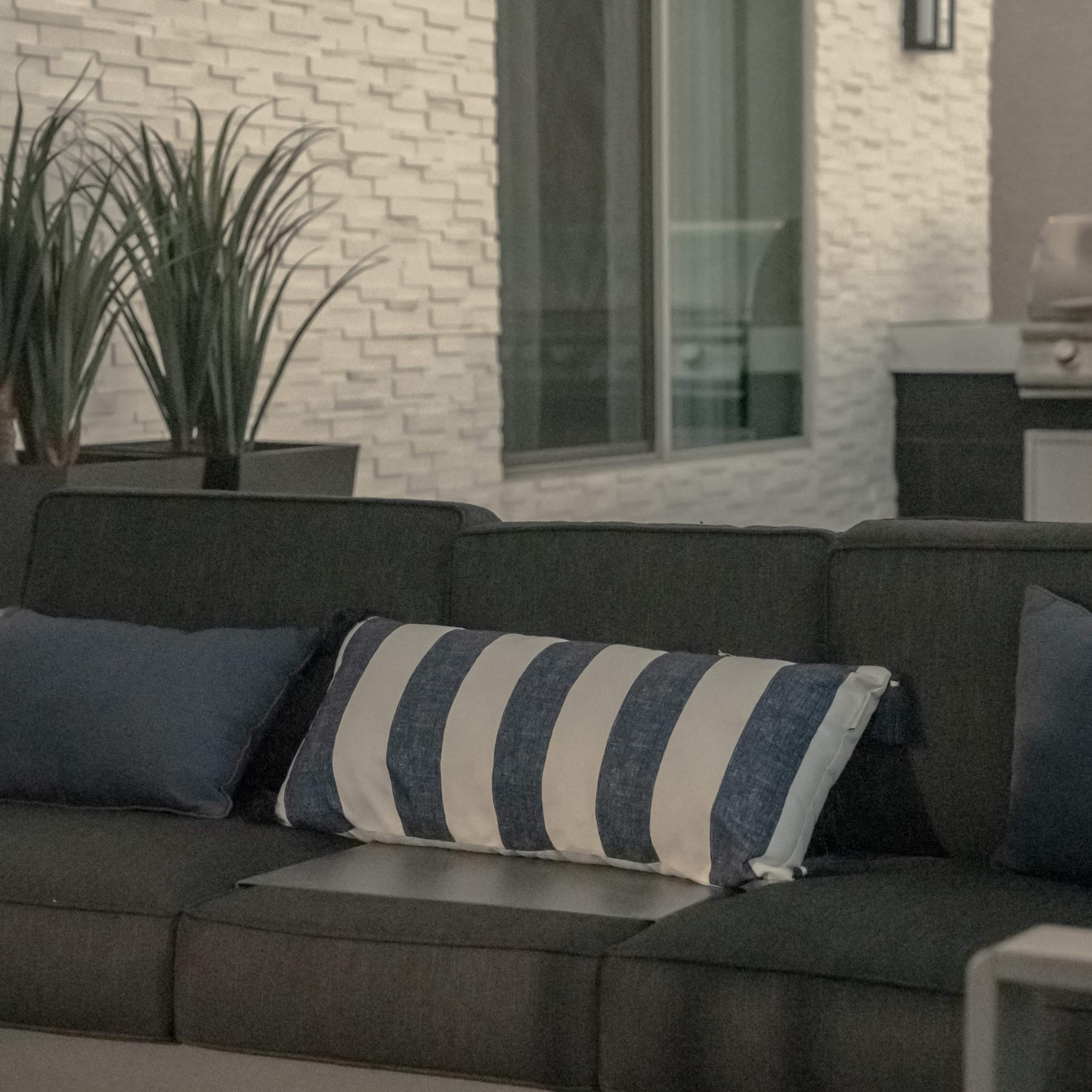 A modern outdoor seating area with a dark gray couch adorned with a striped pillow, beside potted plants and a stainless steel grill in the background.