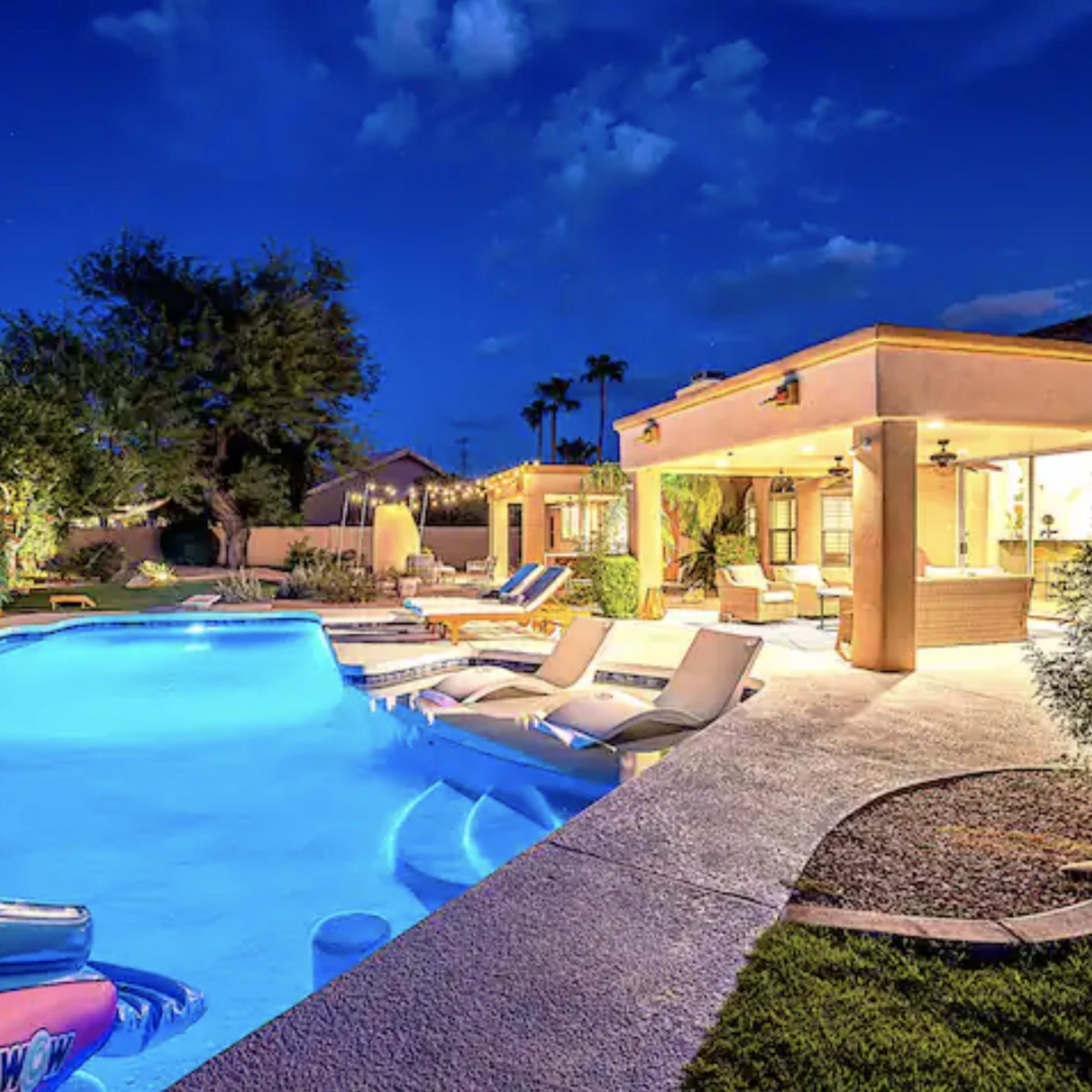 A well-lit backyard features a swimming pool with blue water, surrounded by lounge chairs and landscaping under a night sky.