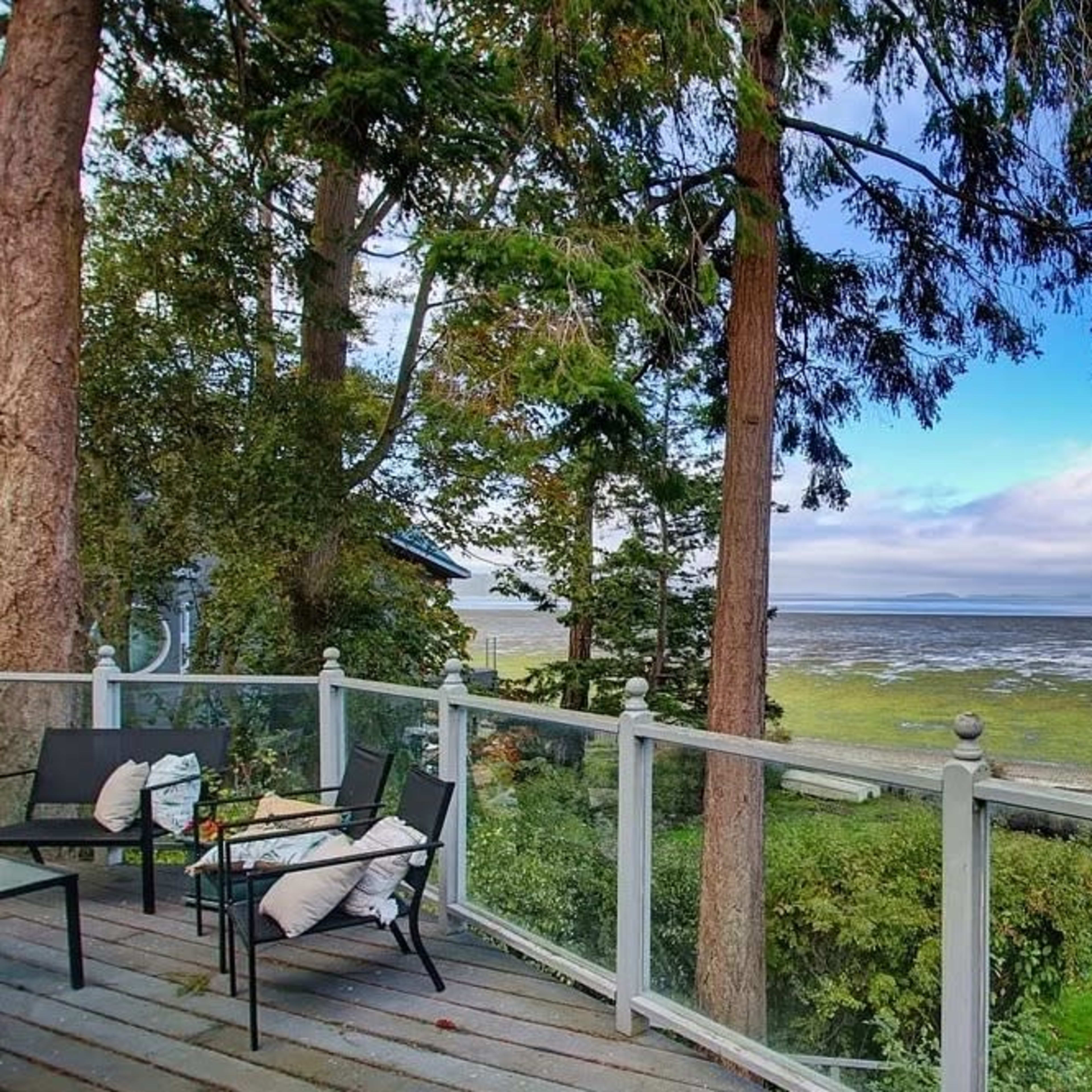 A wooden deck with chairs overlooks a tidal bay surrounded by trees and low vegetation under a partly cloudy sky.