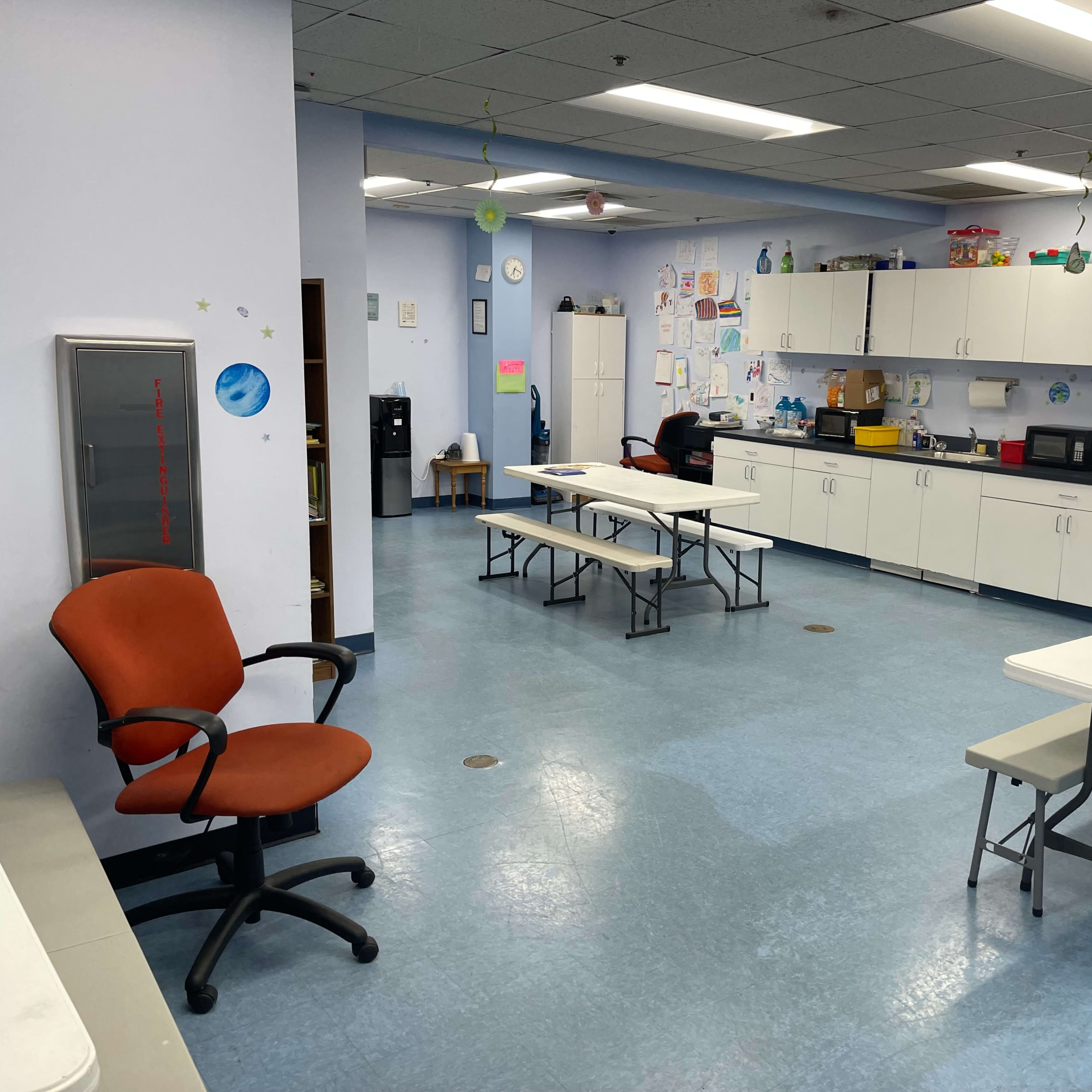 A kitchen or break room with several tables and chairs arranged around the space, and cabinets and appliances along the walls.