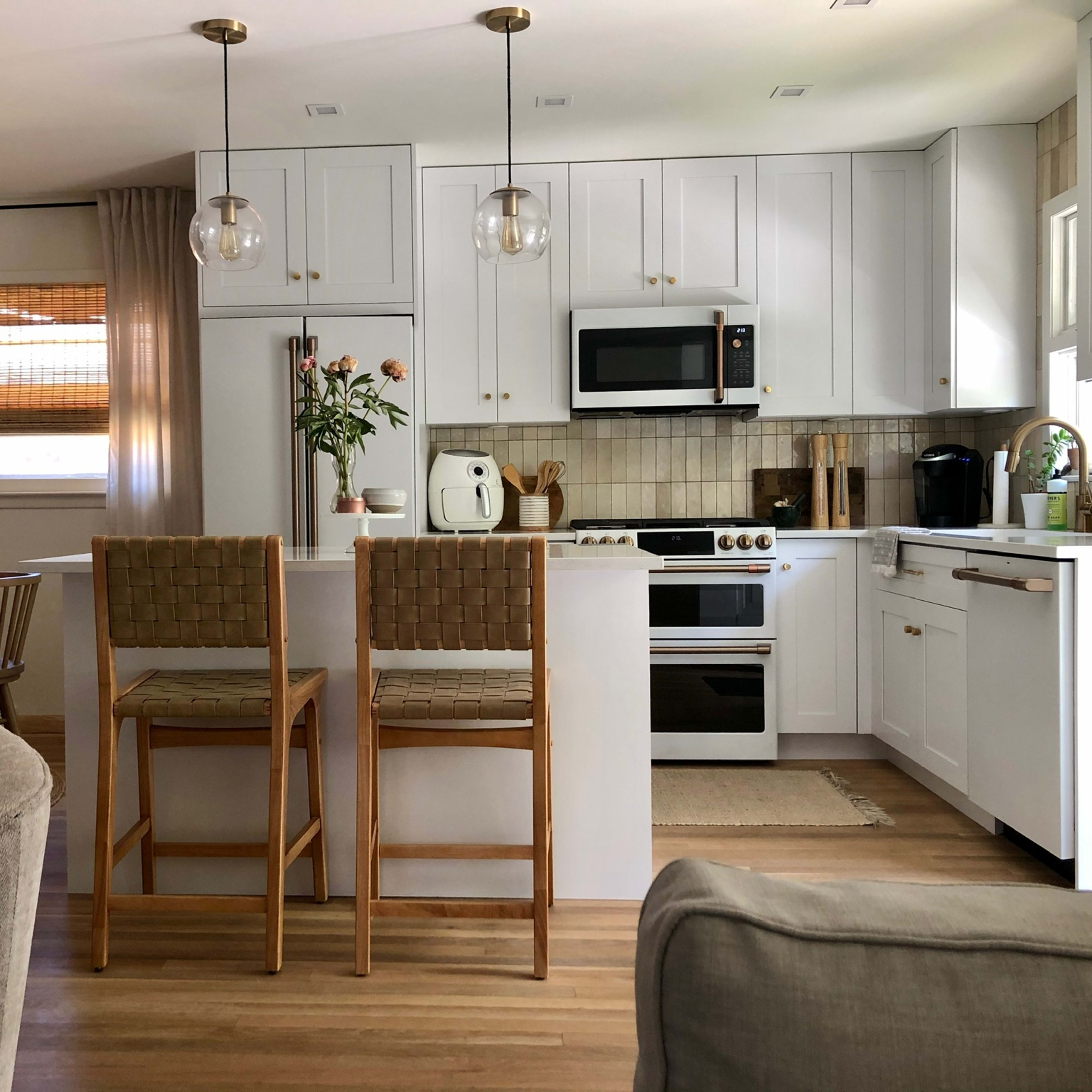A modern kitchen featuring white cabinets, stainless steel appliances, and two barstools at a small island, with a dining area visible in the background.
