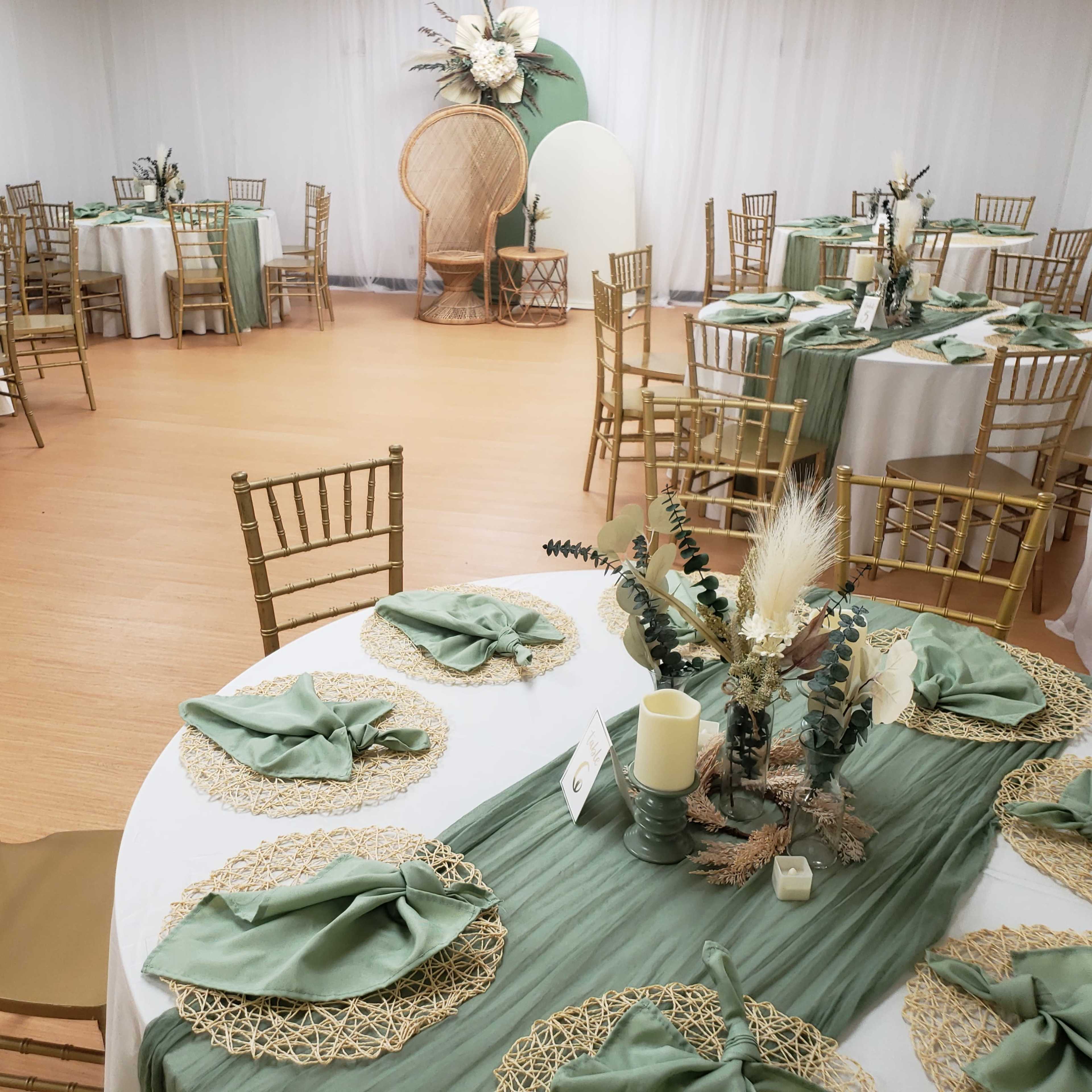 A spacious banquet room is set up with round tables adorned with green table runners, gold accents, and floral decorations, while additional tables are arranged in the background.