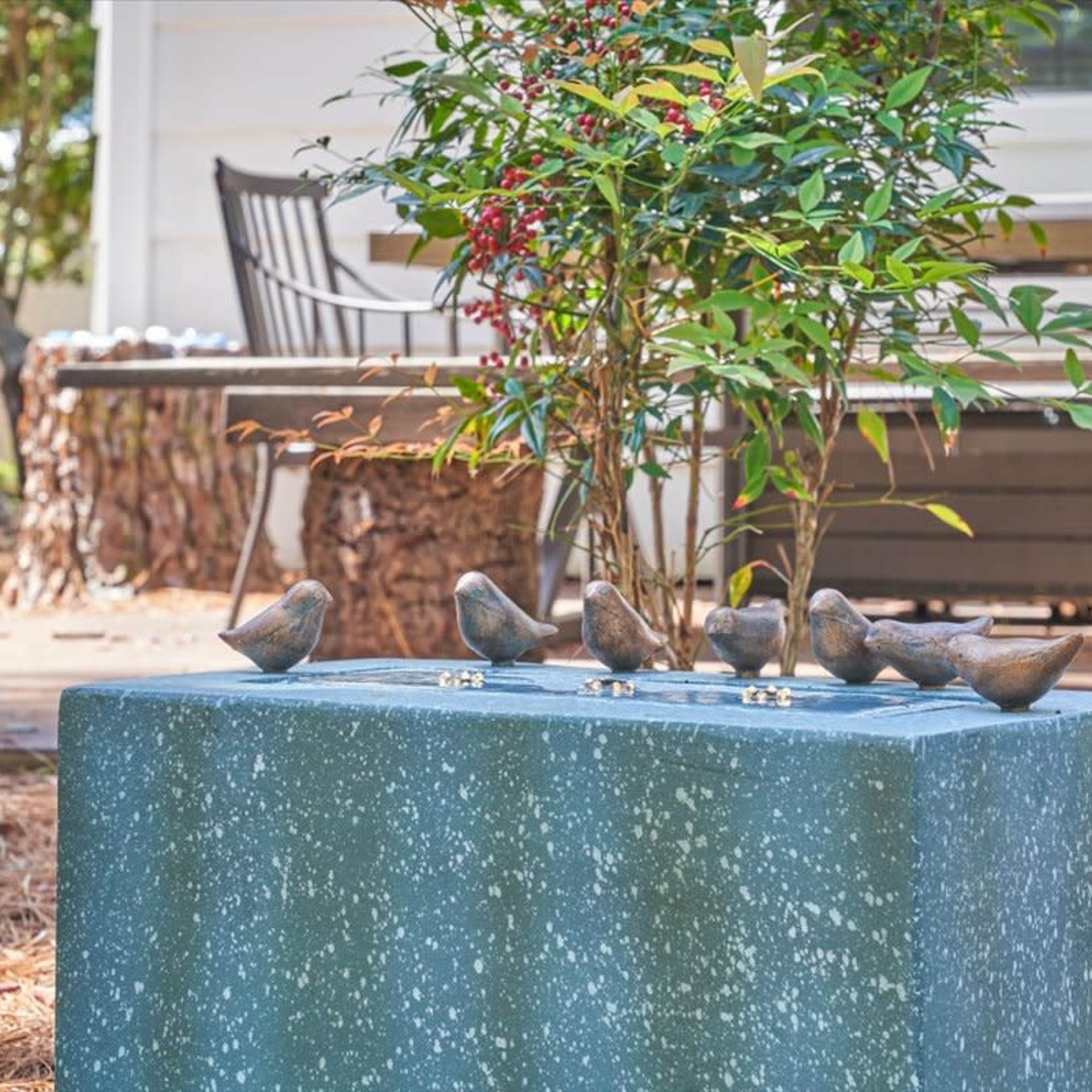A stone pedestal with five bronze bird sculptures is surrounded by green foliage and backed by a wooden bench.