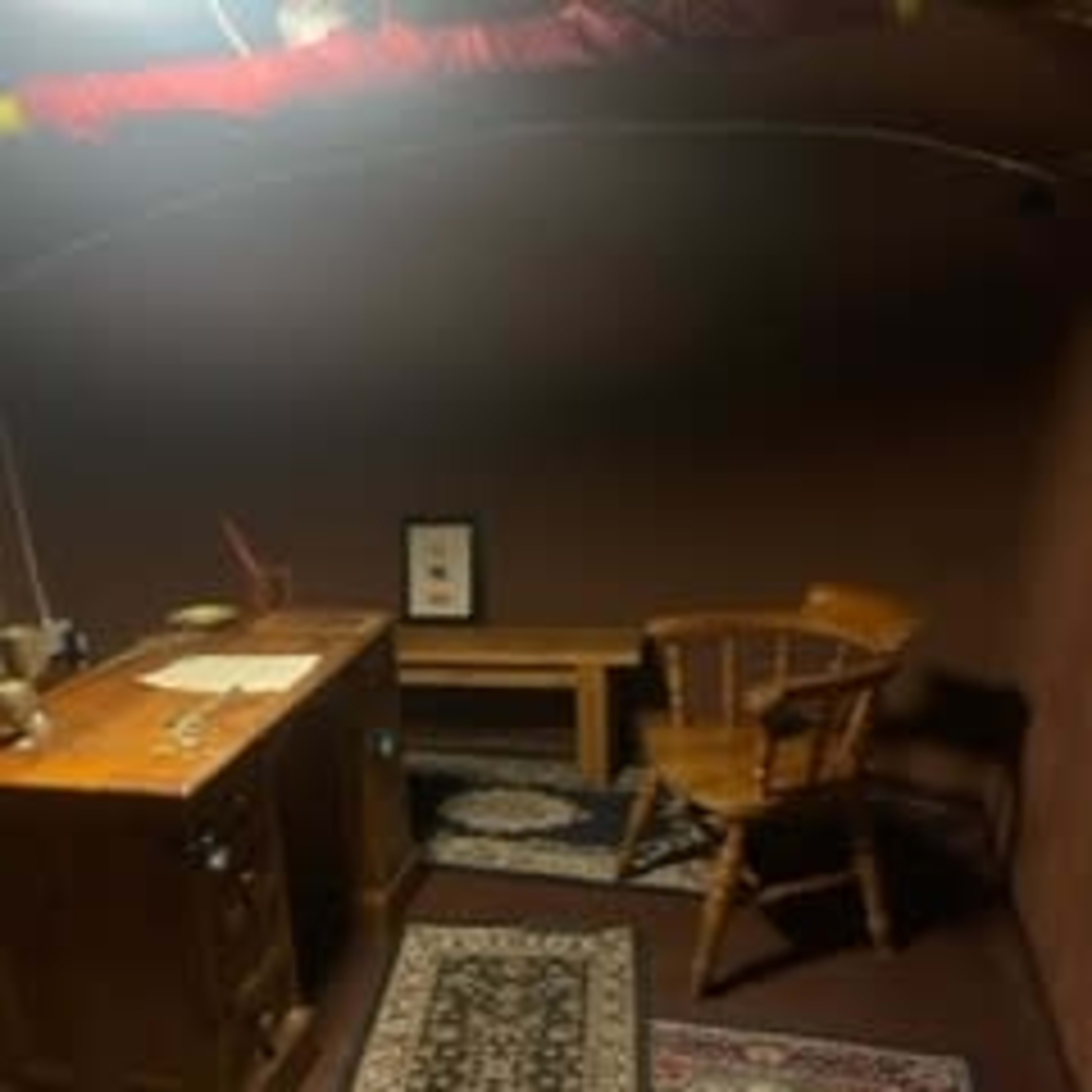 A sparsely furnished room with a wooden desk, a chair, and rugs on the floor.