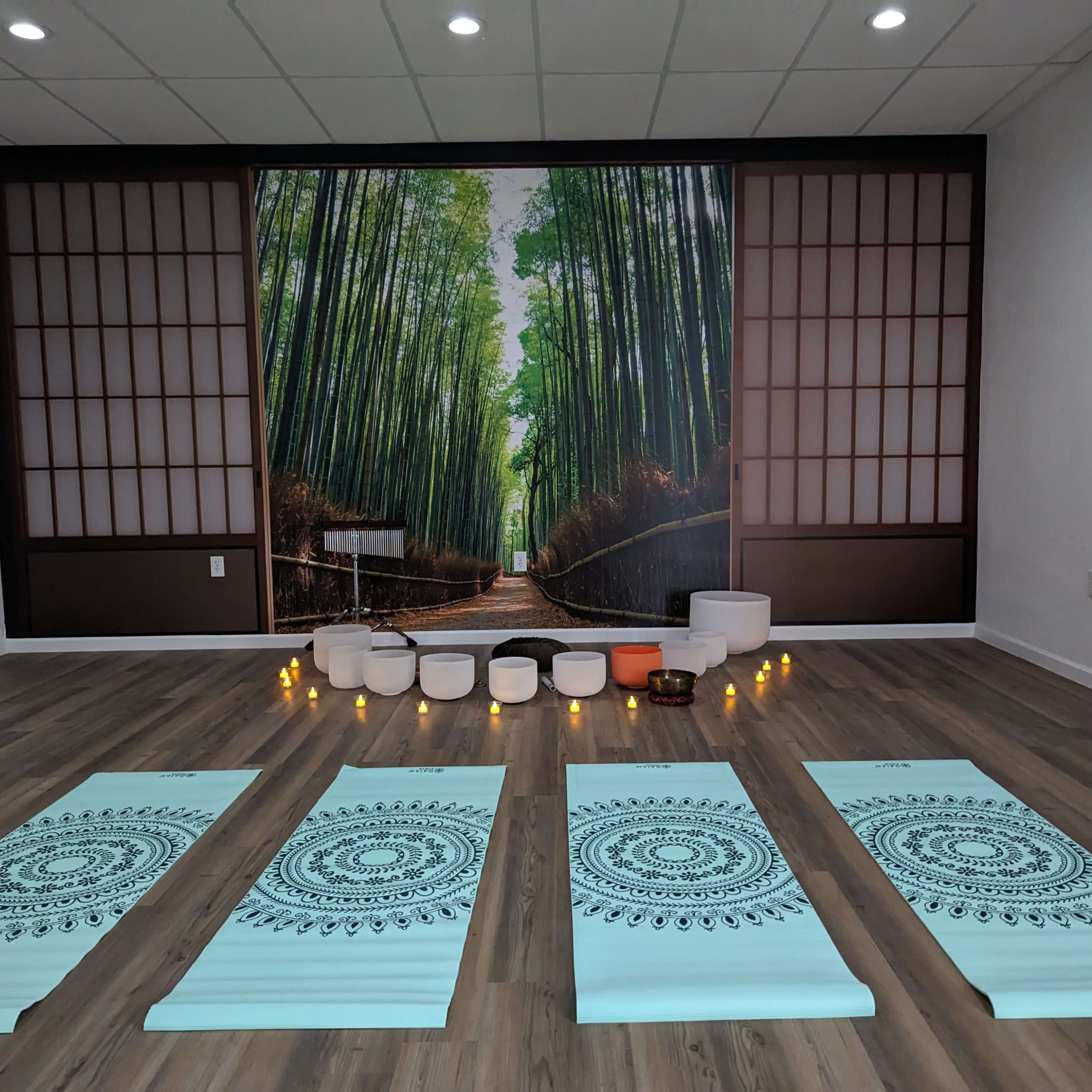 A yoga studio featuring four yoga mats arranged in a row, with a backdrop of a forest mural and bowls set up at the front, surrounded by candles.