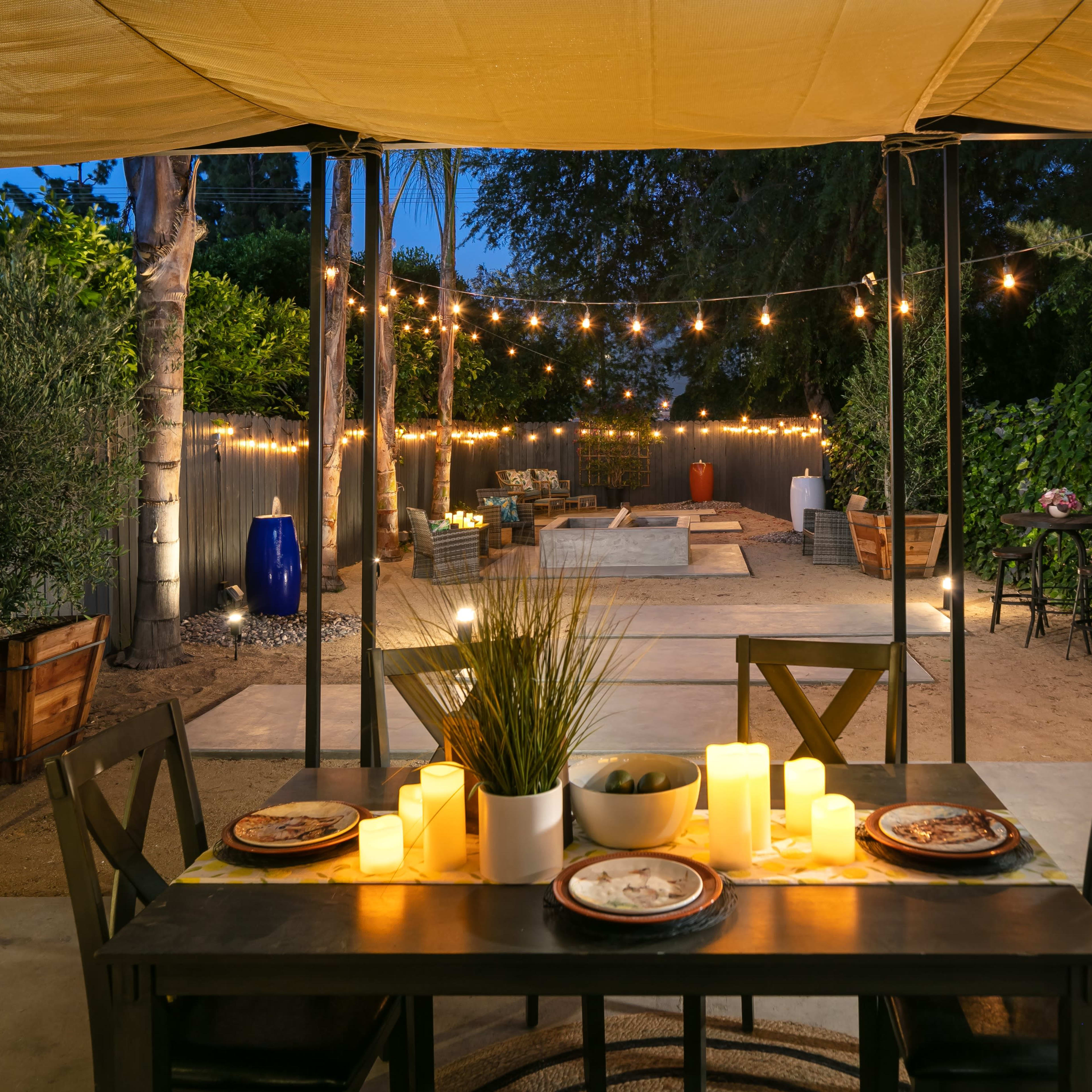A table set with plates and lit candles is positioned under a canopy, overlooking a backyard adorned with string lights and potted plants.