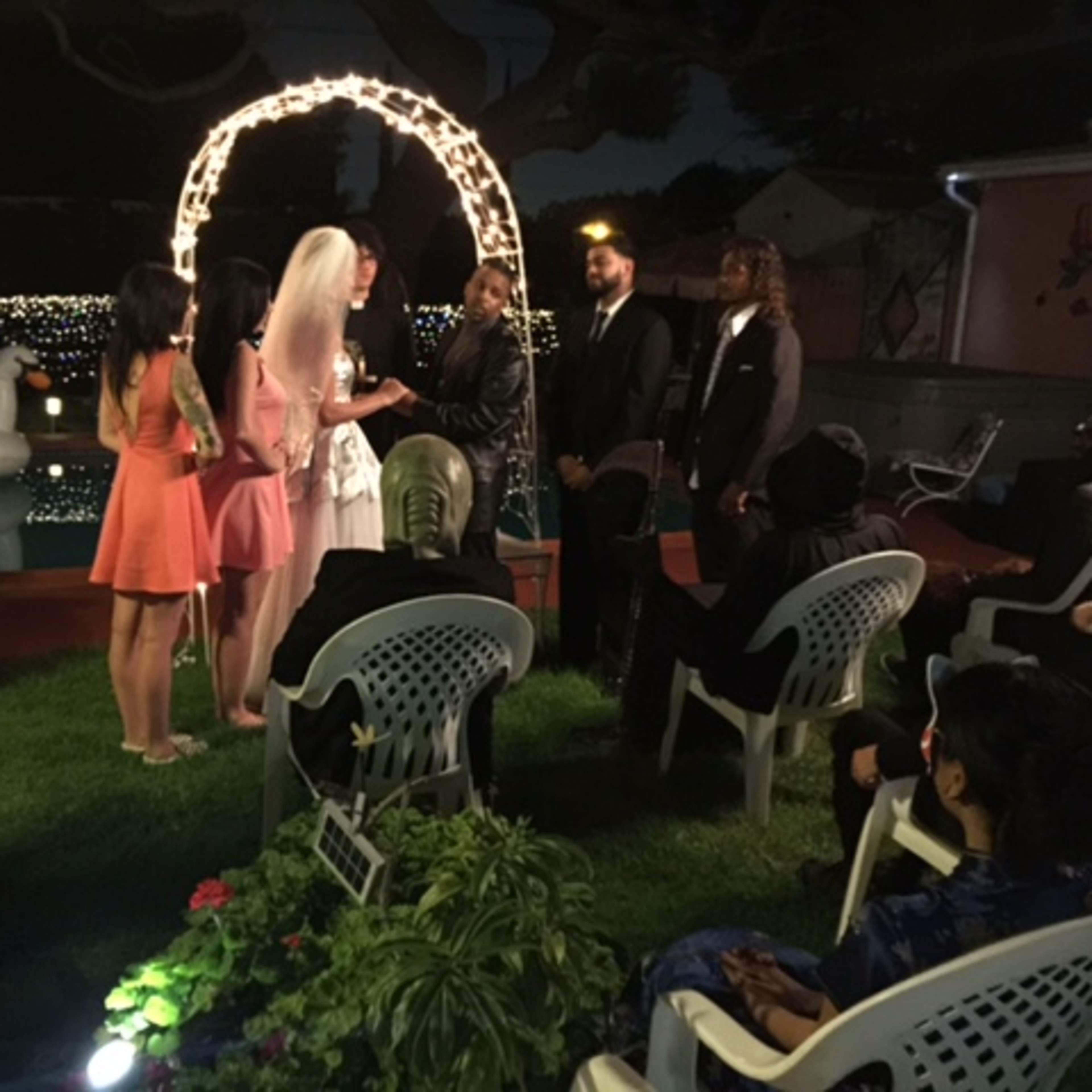 A couple exchanges vows under a lighted arch in a backyard during a nighttime wedding ceremony.