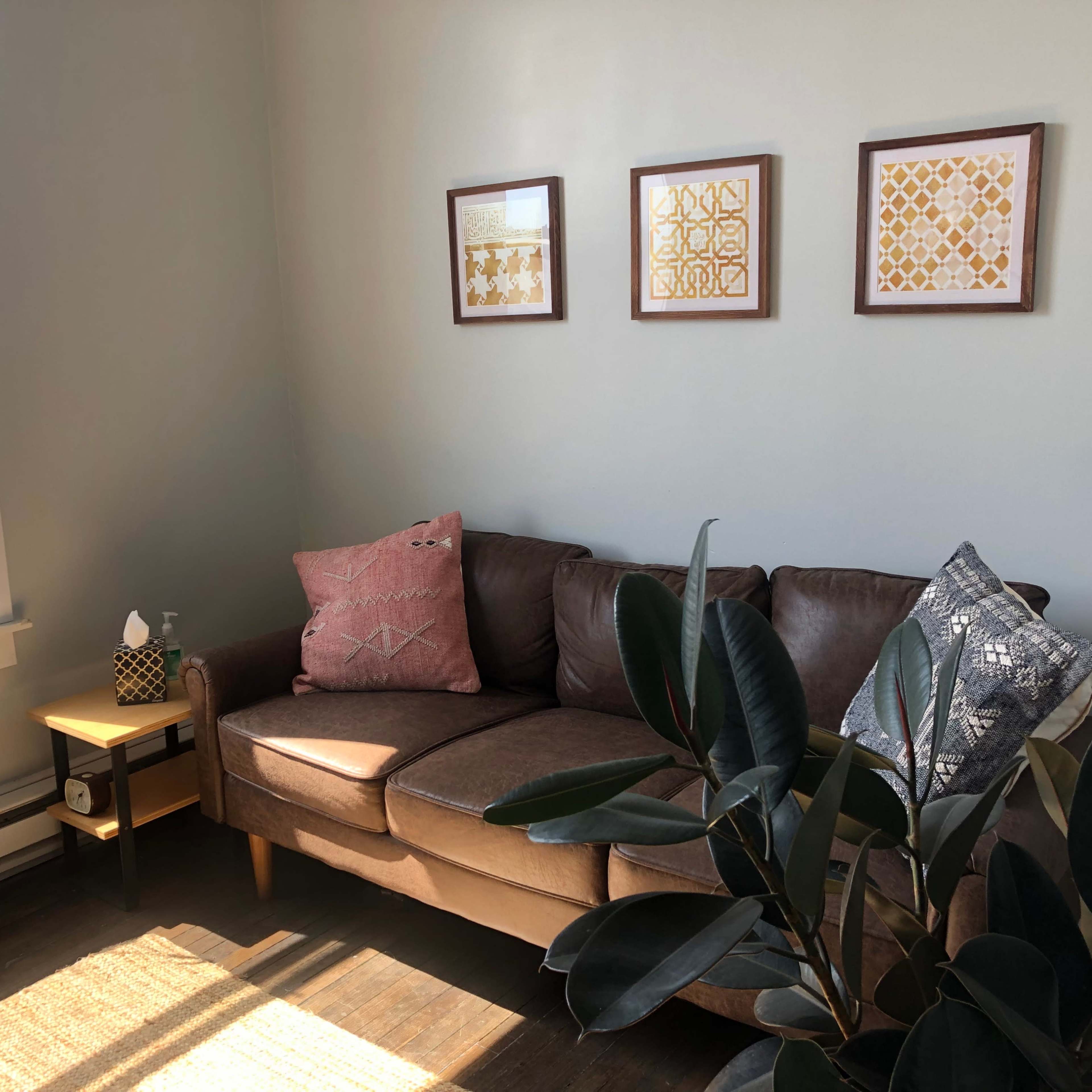 A brown couch with decorative pillows sits in a bright corner of a room, accompanied by a small side table and three framed artworks on the wall.