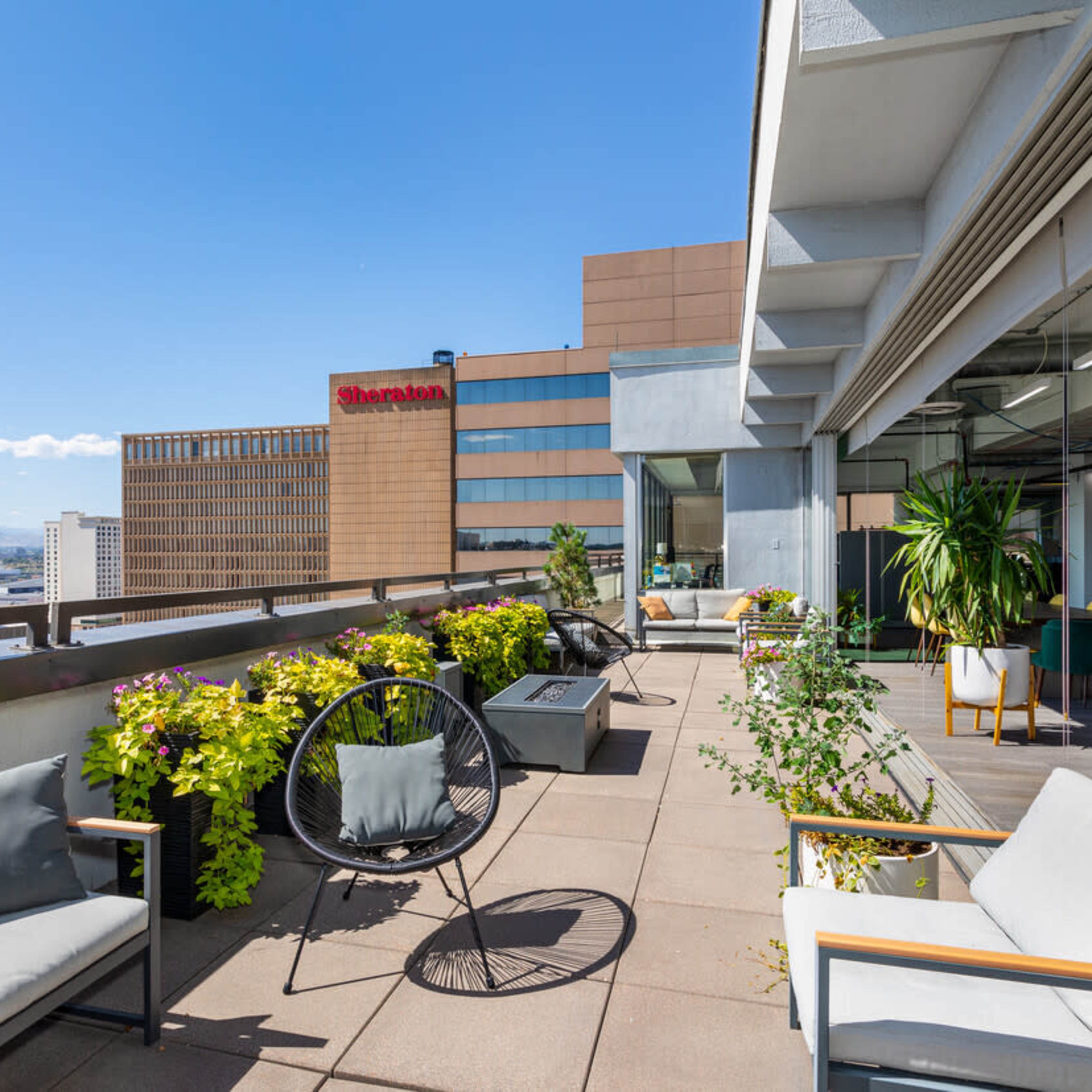 A rooftop patio with seating, potted plants, and a view of buildings including a Sheraton hotel in the background.