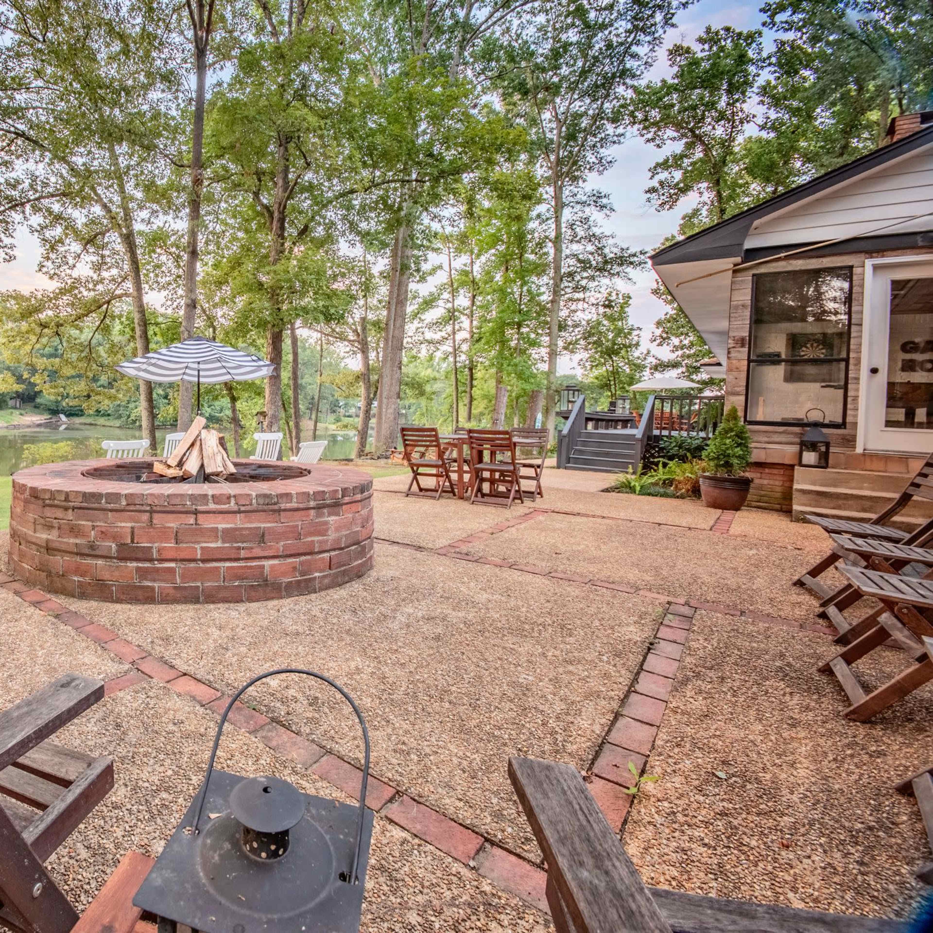 A circular stone fire pit surrounded by wooden chairs and a picnic table overlooks a calm lake through a grove of trees.