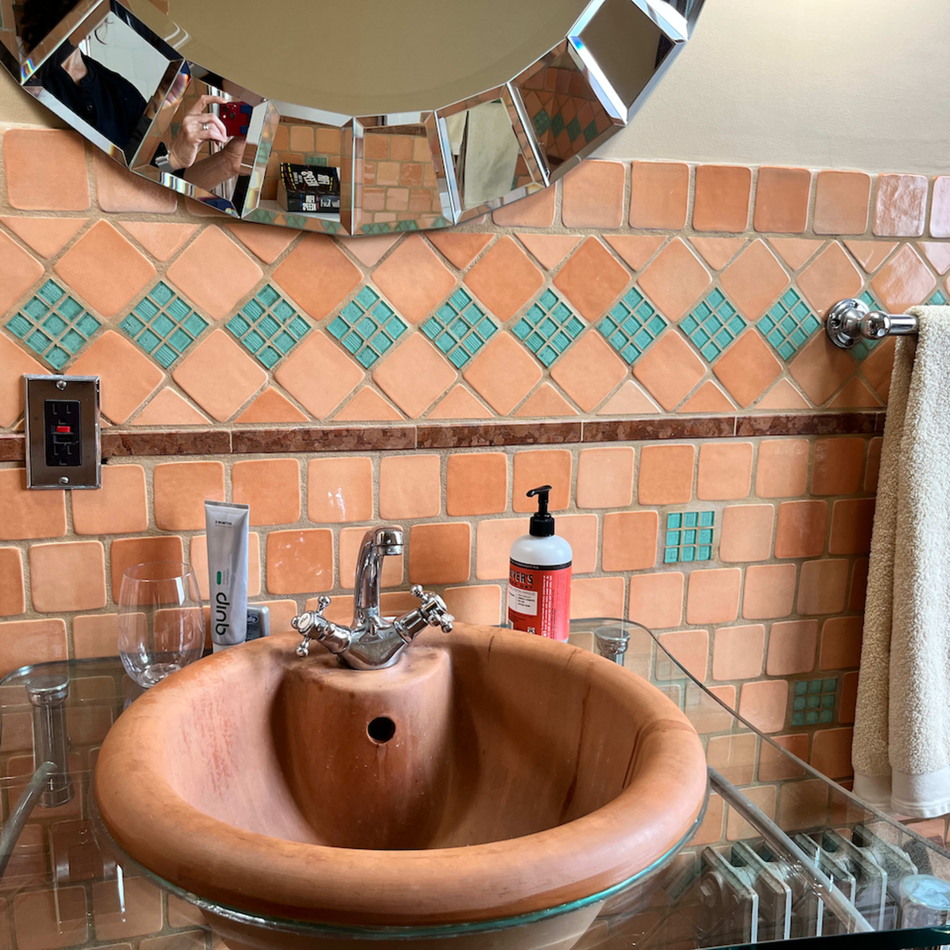 A bathroom sink made of terracotta, surrounded by tiled walls in orange and green, with a mirror reflecting the space.