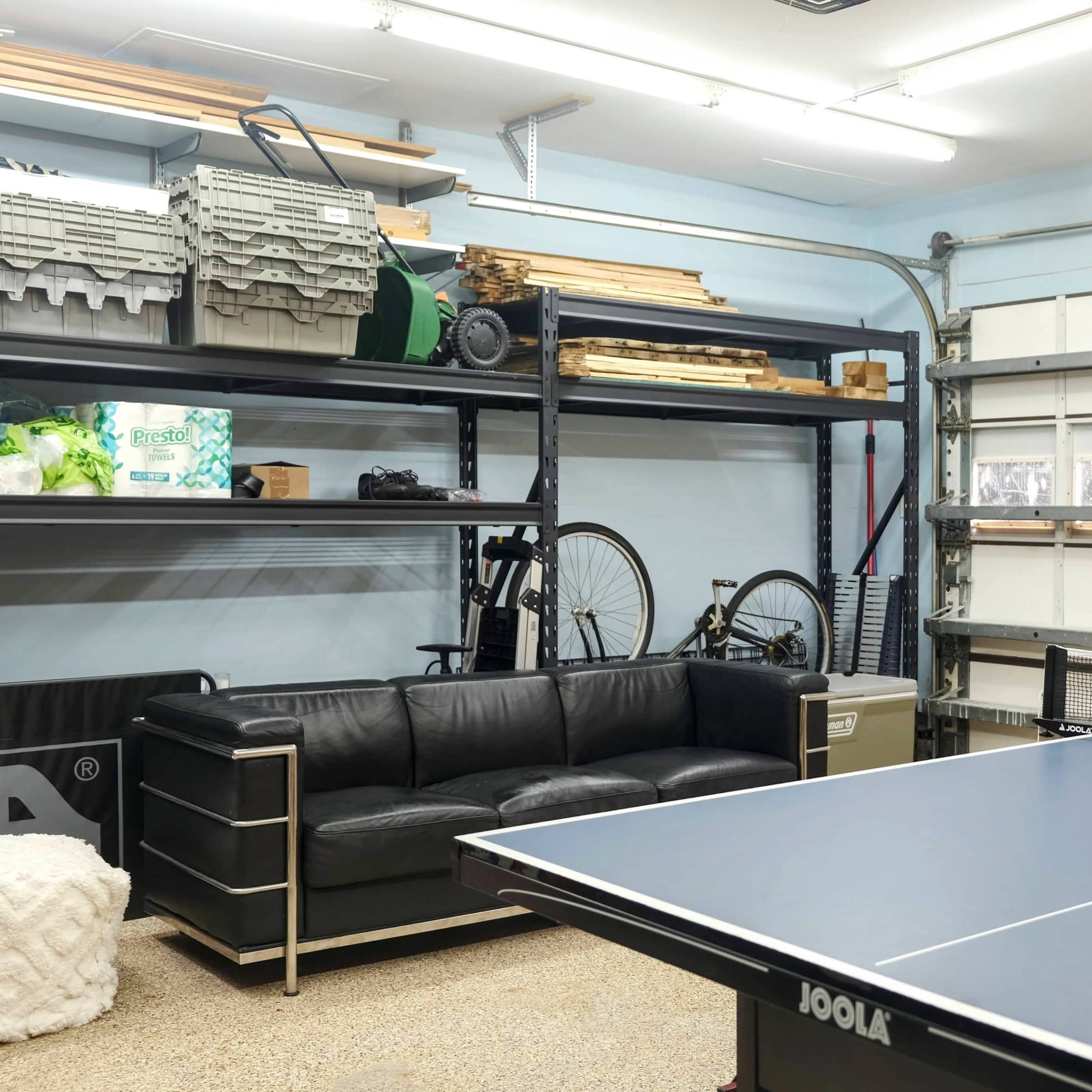 A garage equipped with a ping pong table, a black couch, shelving units filled with storage bins, and bicycles in the background.