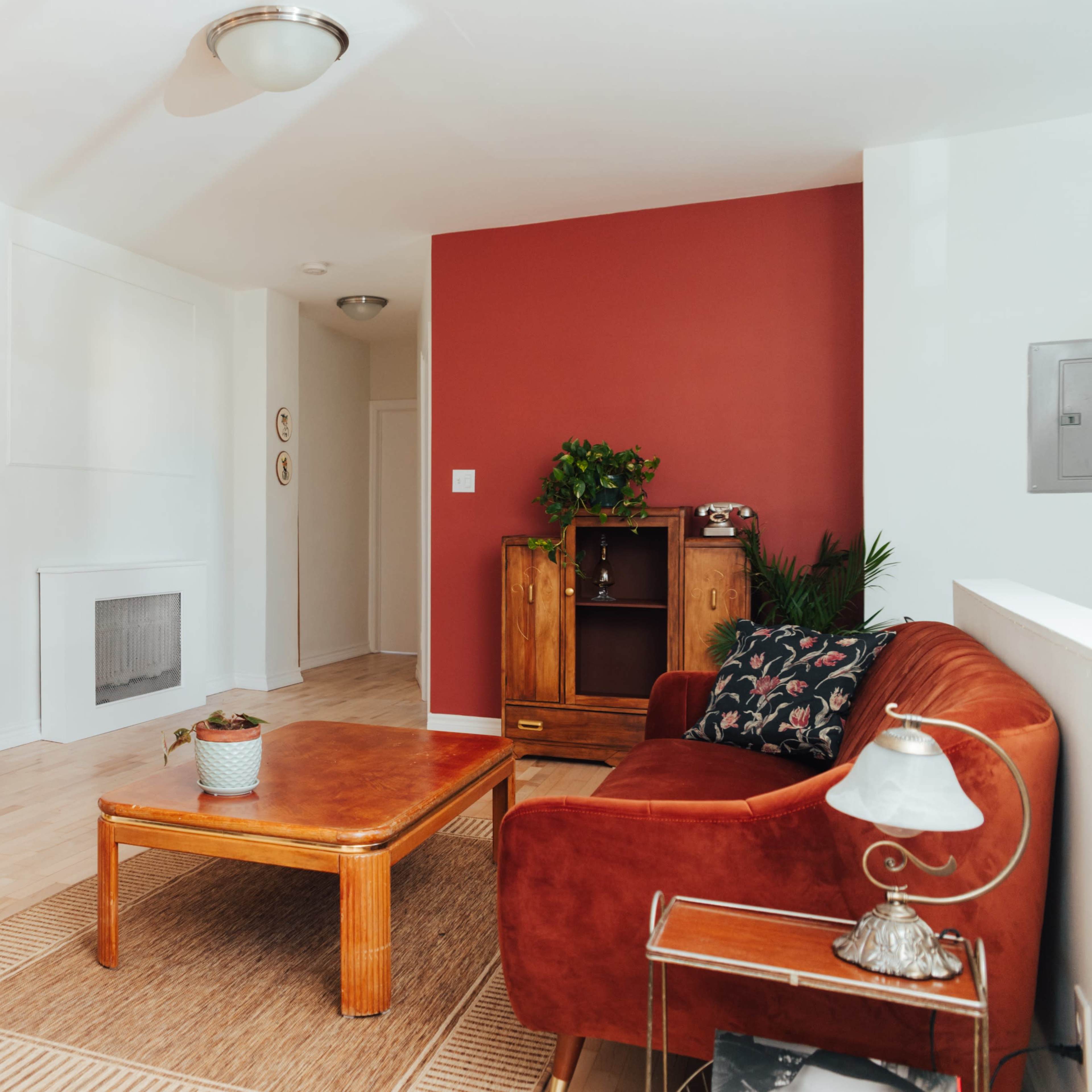 The room features a wooden fireplace mantel, a red accent wall, a brown couch, and a coffee table surrounded by plants.