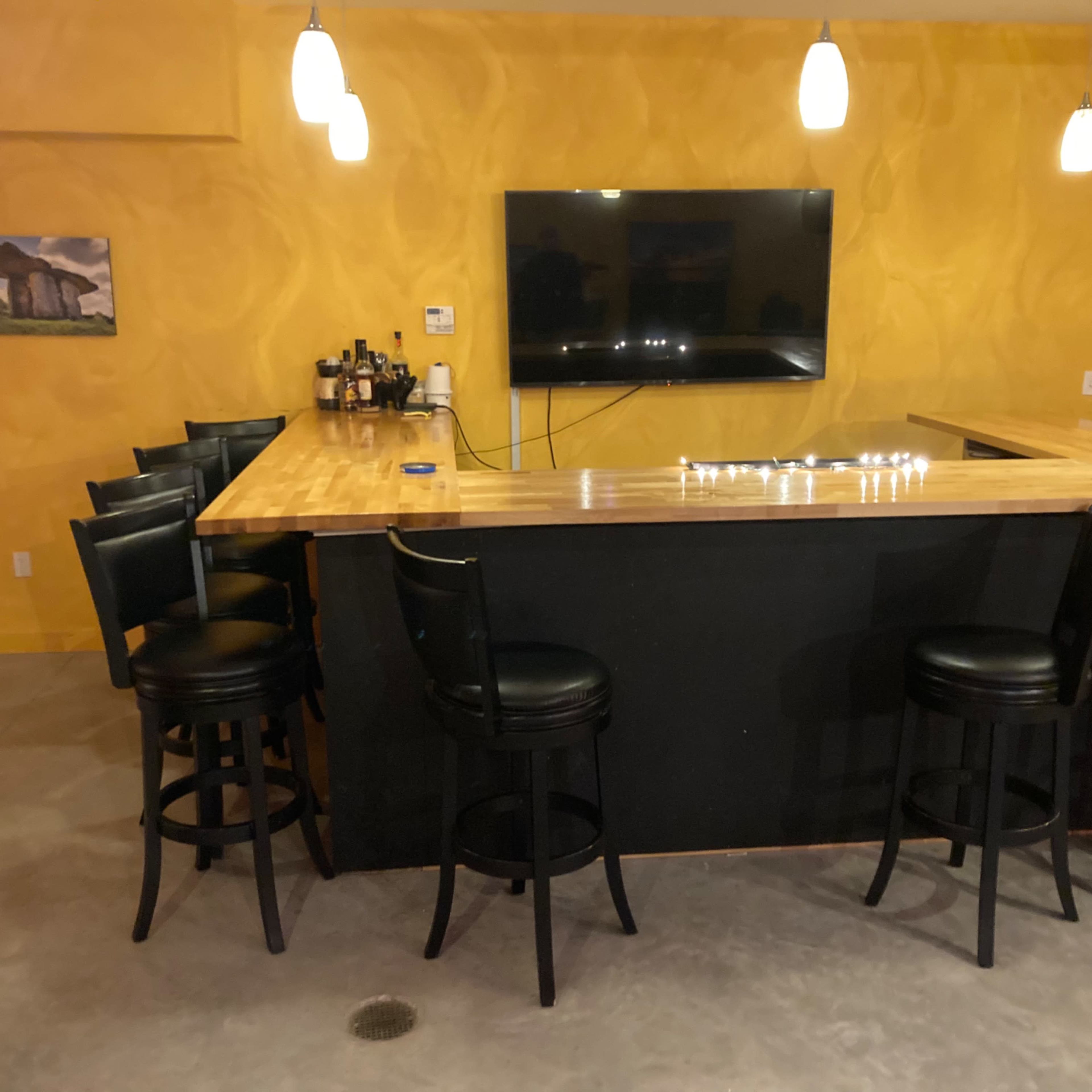 A bar setup with a wooden countertop, six black stools, a mounted television, and a yellow wall in a dimly lit room.