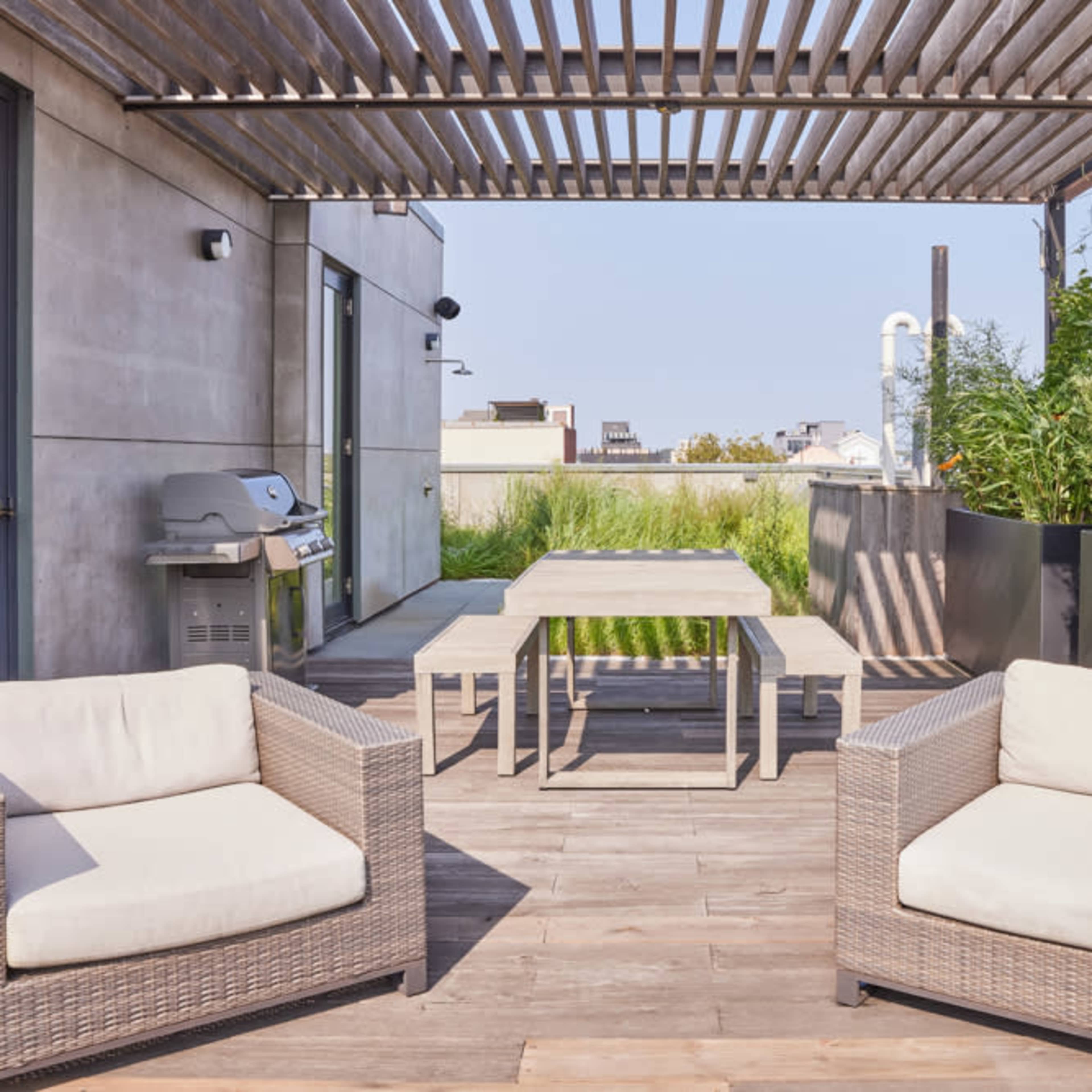 A modern rooftop terrace featuring two wicker armchairs, a wooden table, and a barbecue grill under a slatted pergola.