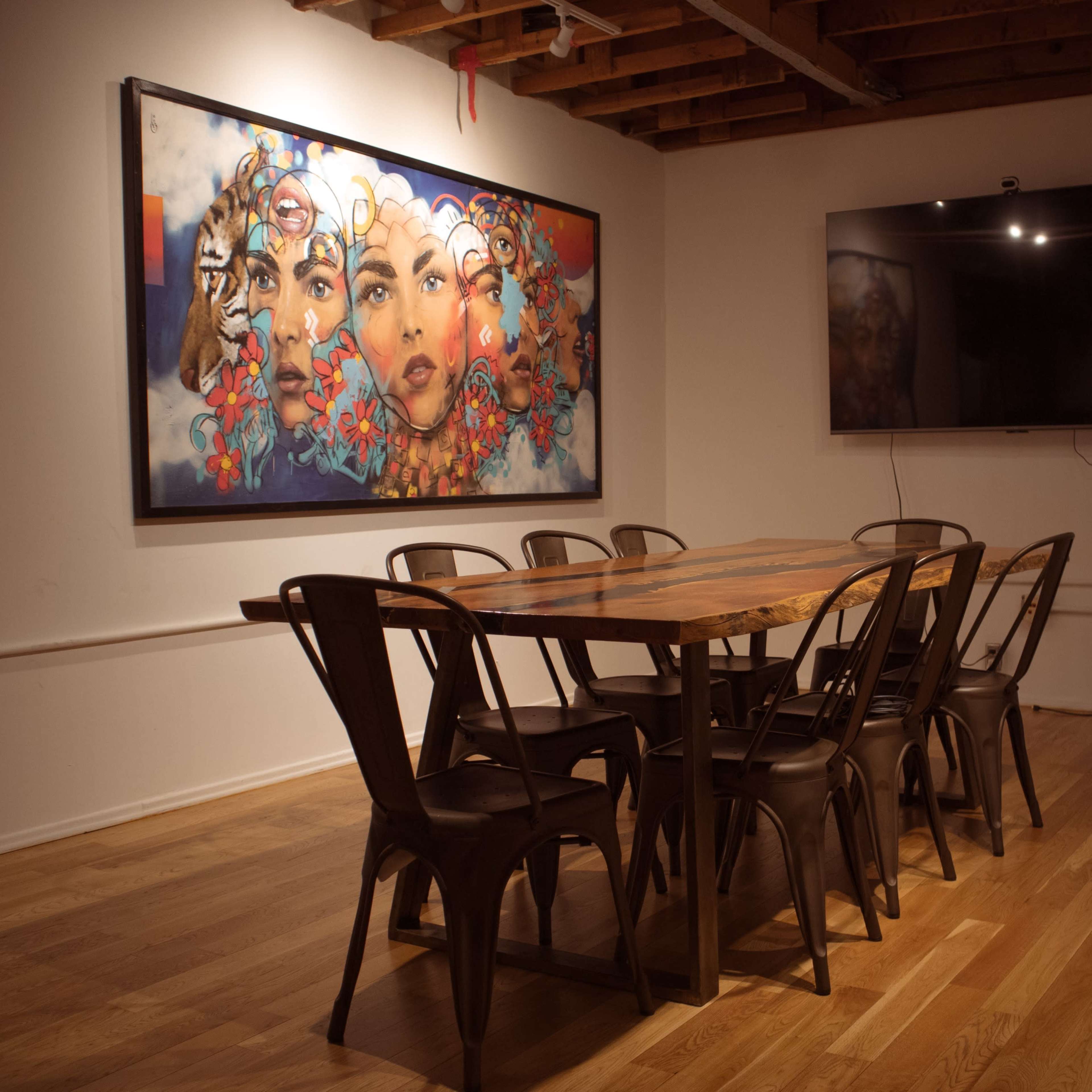 A wooden dining table with eight metal chairs is set in a room featuring a large colorful mural on one wall and a flat-screen TV on another.
