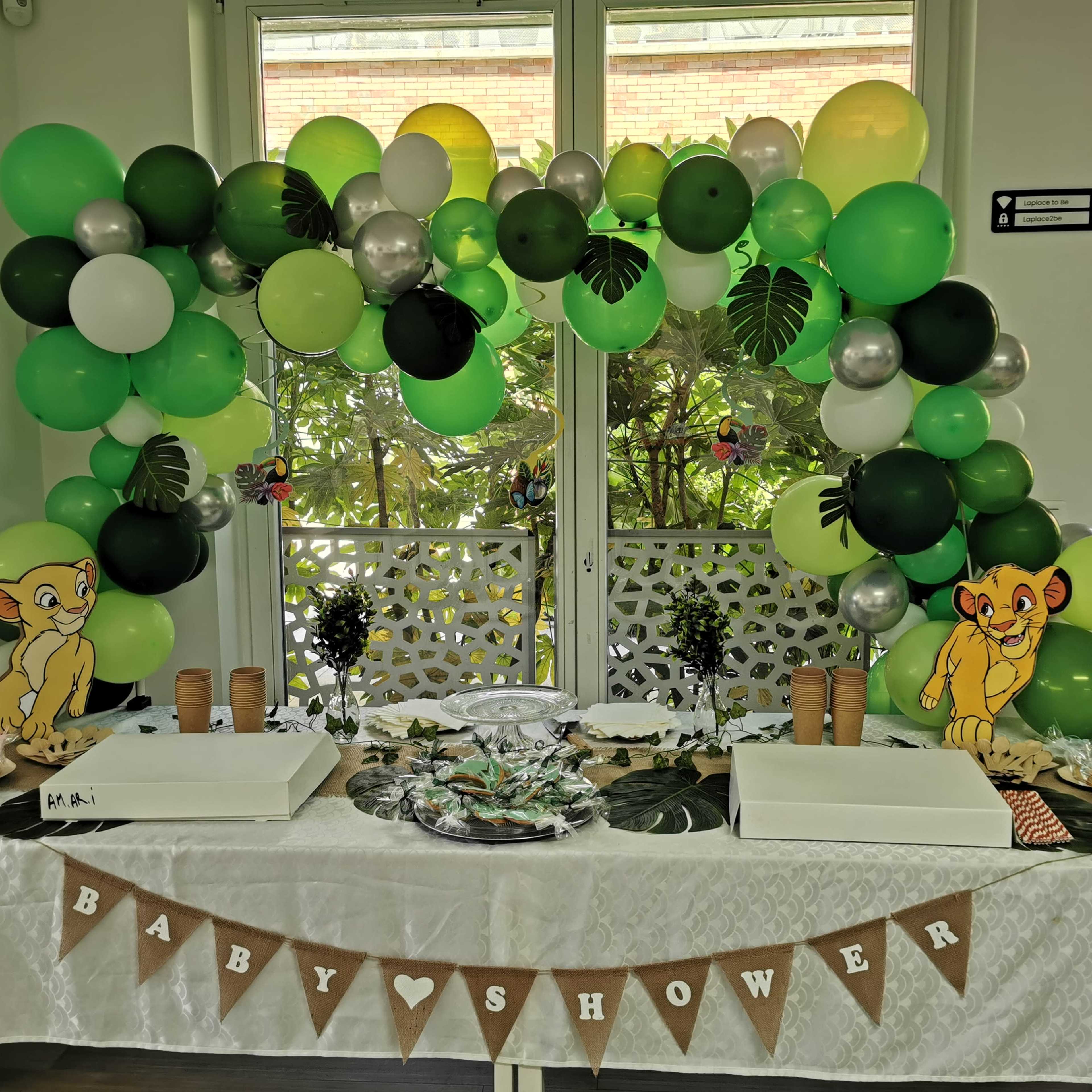 A baby shower table is decorated with green and black balloons, featuring a jungle theme with cartoon lion characters and a "BABY SHOWER" banner.