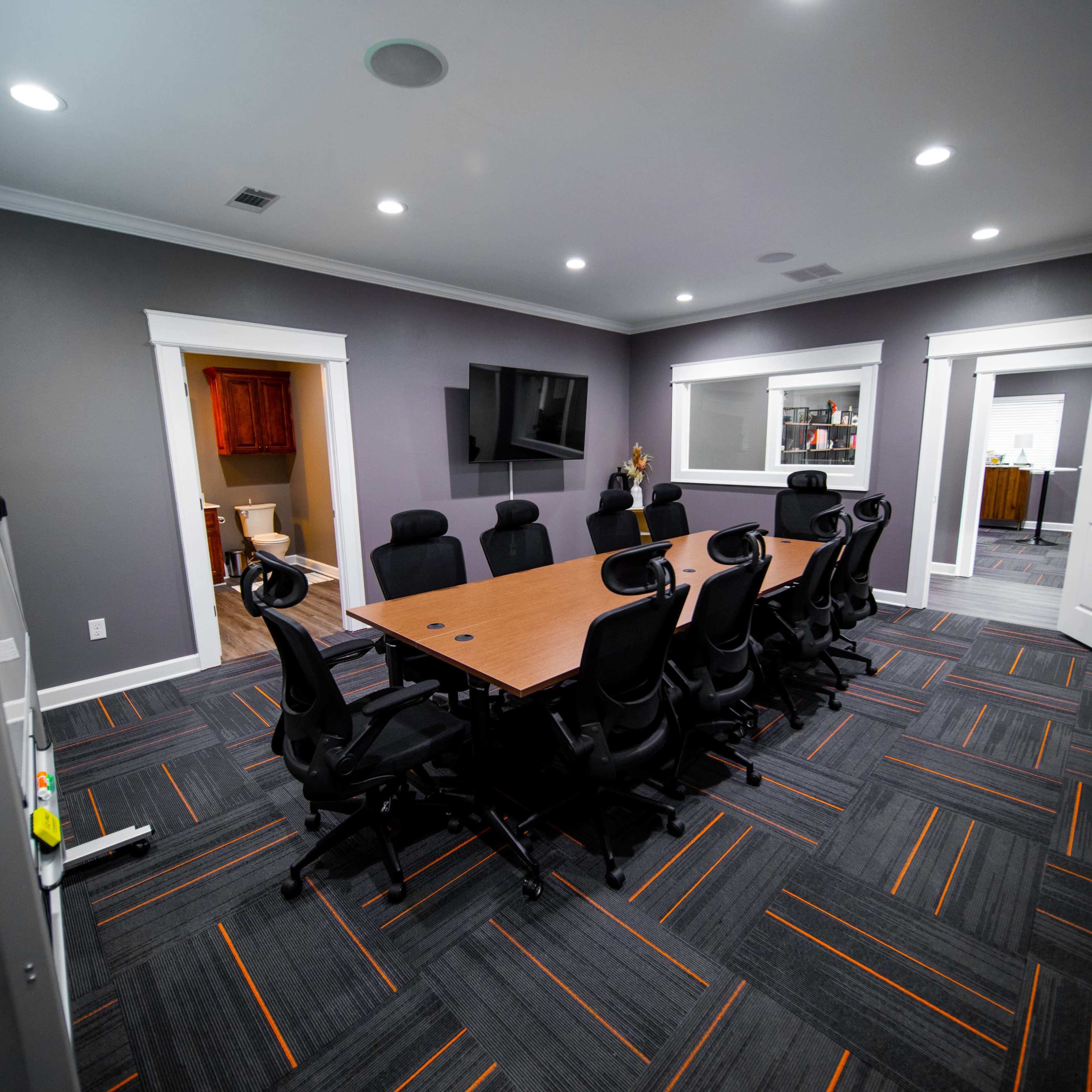 A modern conference room with a large rectangular table surrounded by black office chairs and features two doorways leading to other rooms.